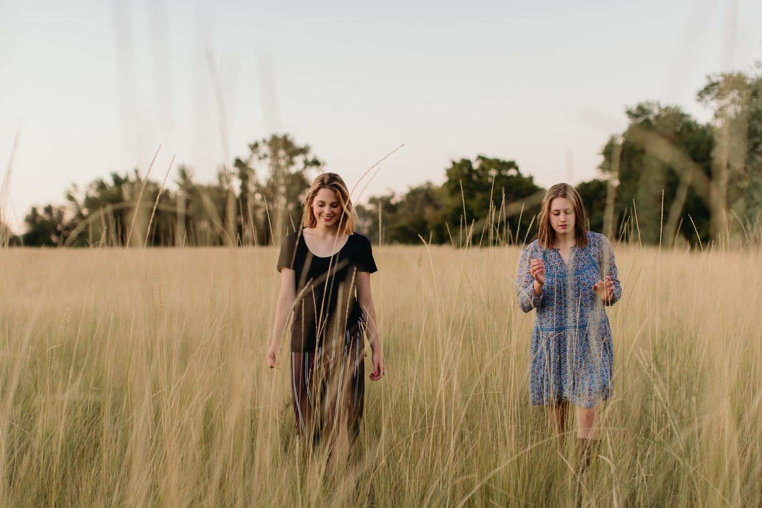 Two women in dresses walking through tall golden grass field at sunset with trees in background.