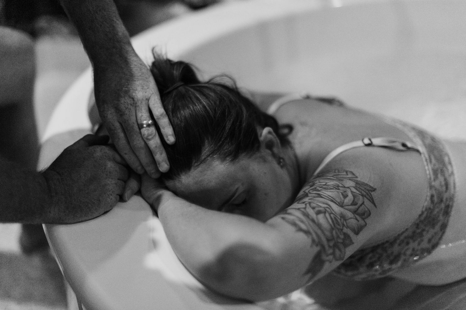 Black and white photo of a tattooed woman leaning over a birthing tub during water labor with supportive hands.