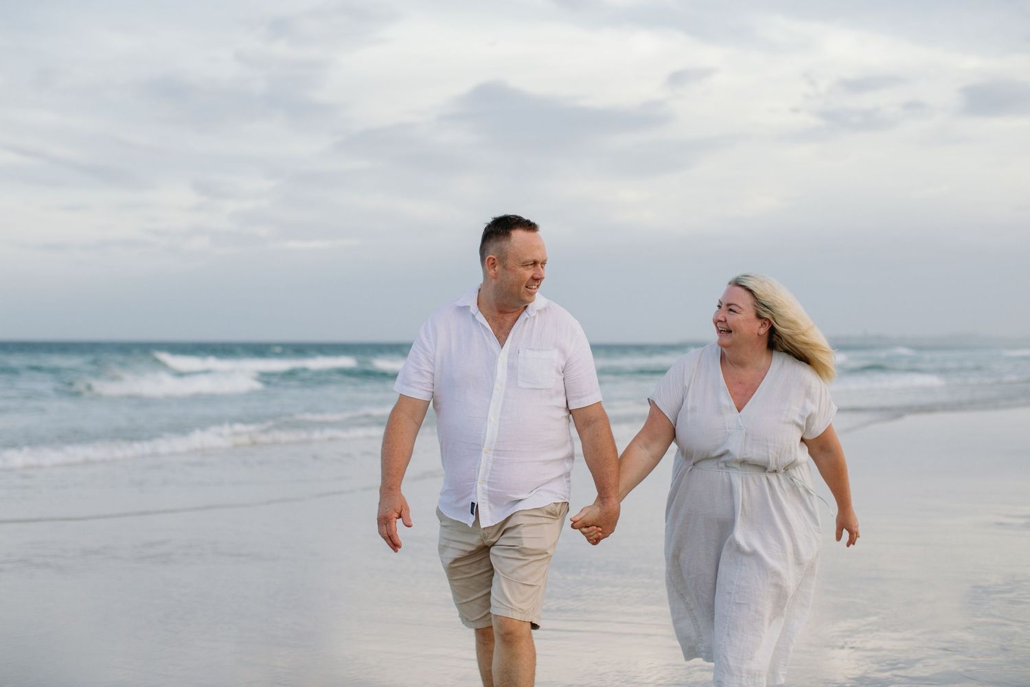 Happy couple in white outfits holding hands and walking along a sandy beach with ocean waves in the background.