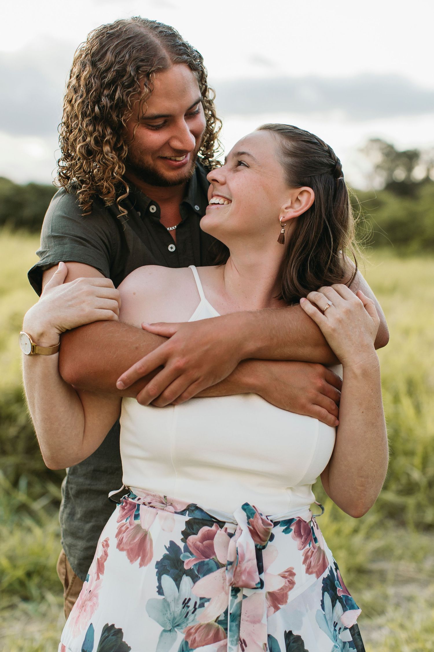 A smiling couple embraces outdoors in a green field, woman in floral skirt and white top, man with curly hair.