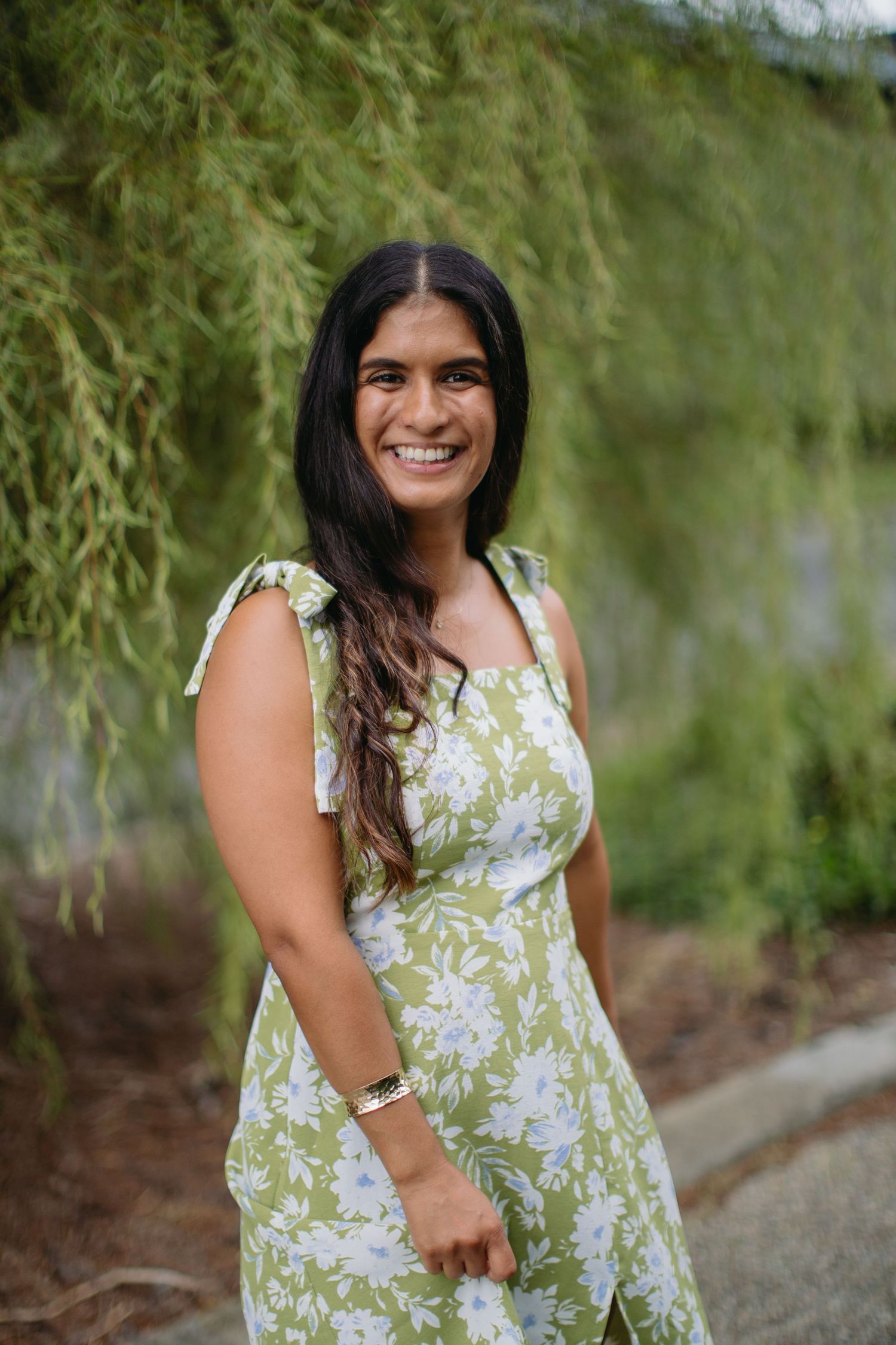 Young woman in green floral sundress smiling outdoors near weeping willow tree branches.