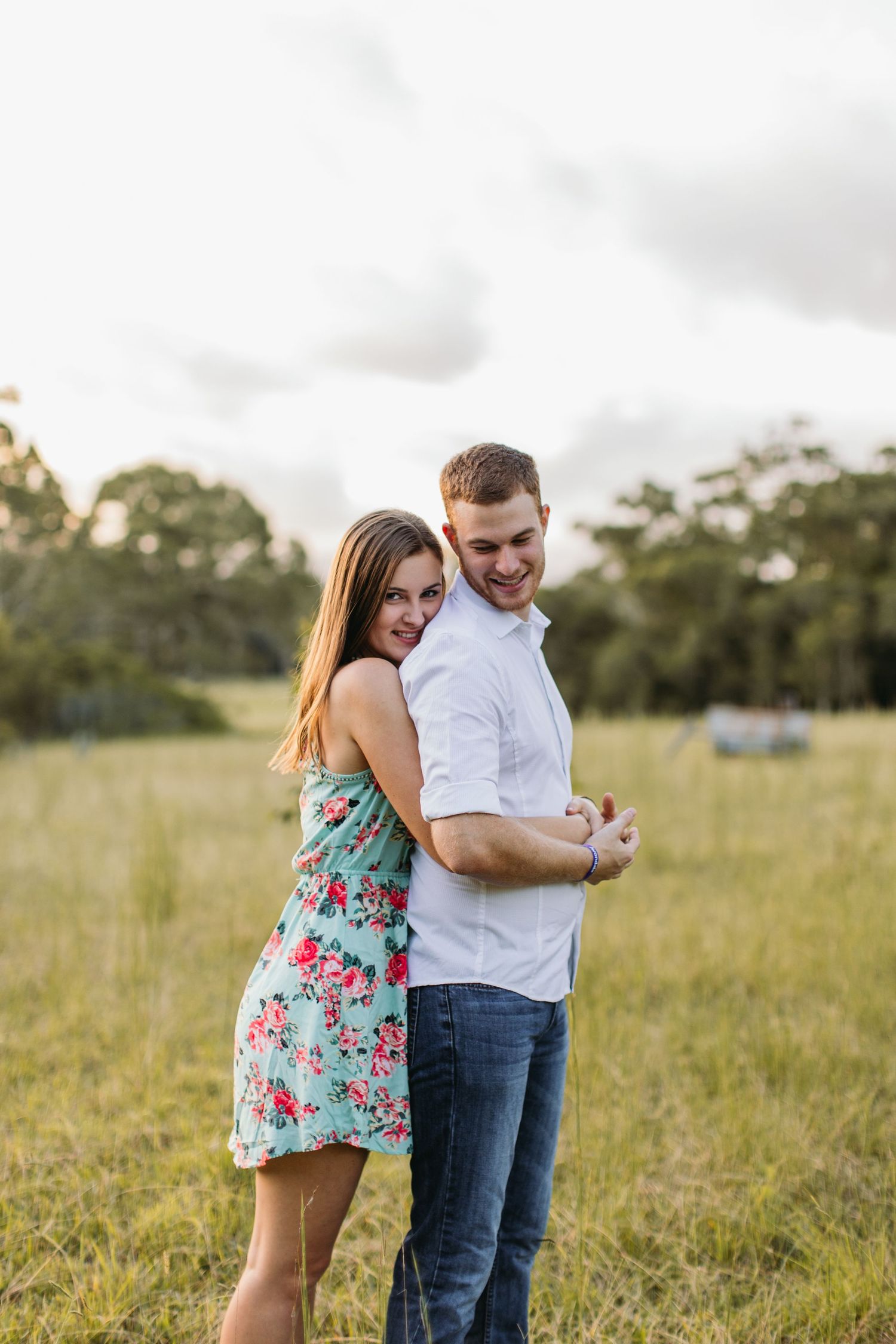 A couple embraces in a green field, woman in floral dress leaning on man in white shirt and jeans.