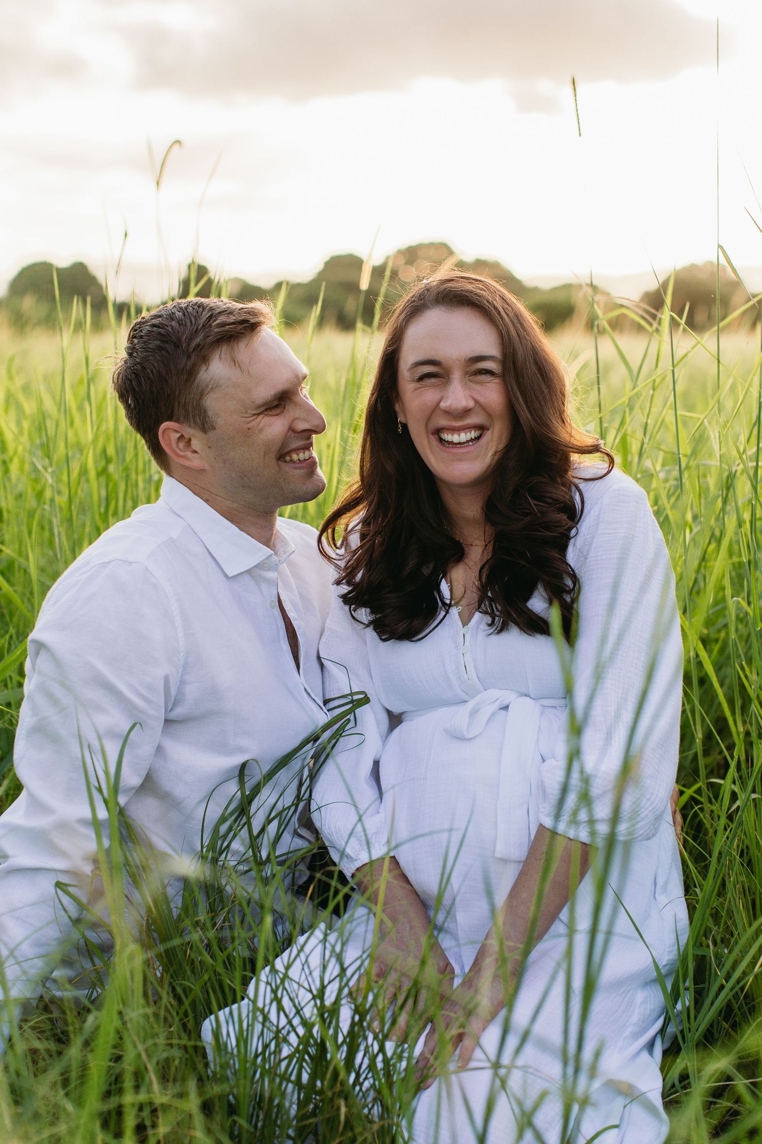 A smiling couple in white outfits sits together in a lush green field at golden hour.