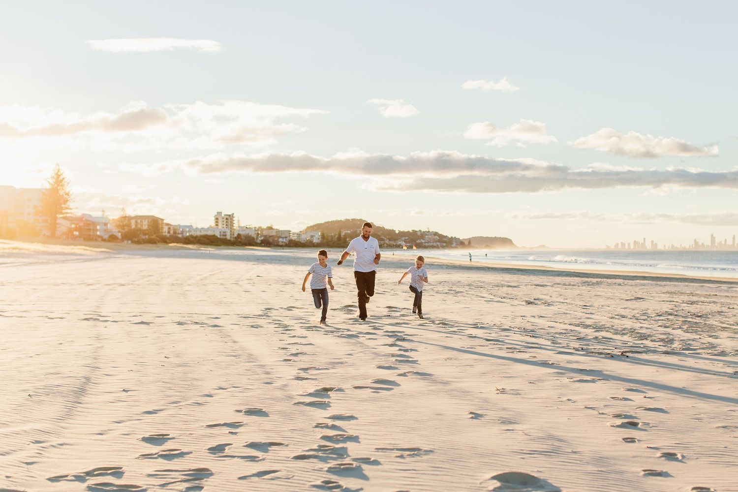 Family walking on a sunny beach at golden hour, leaving footprints in the sand with a coastal cityscape in the background.