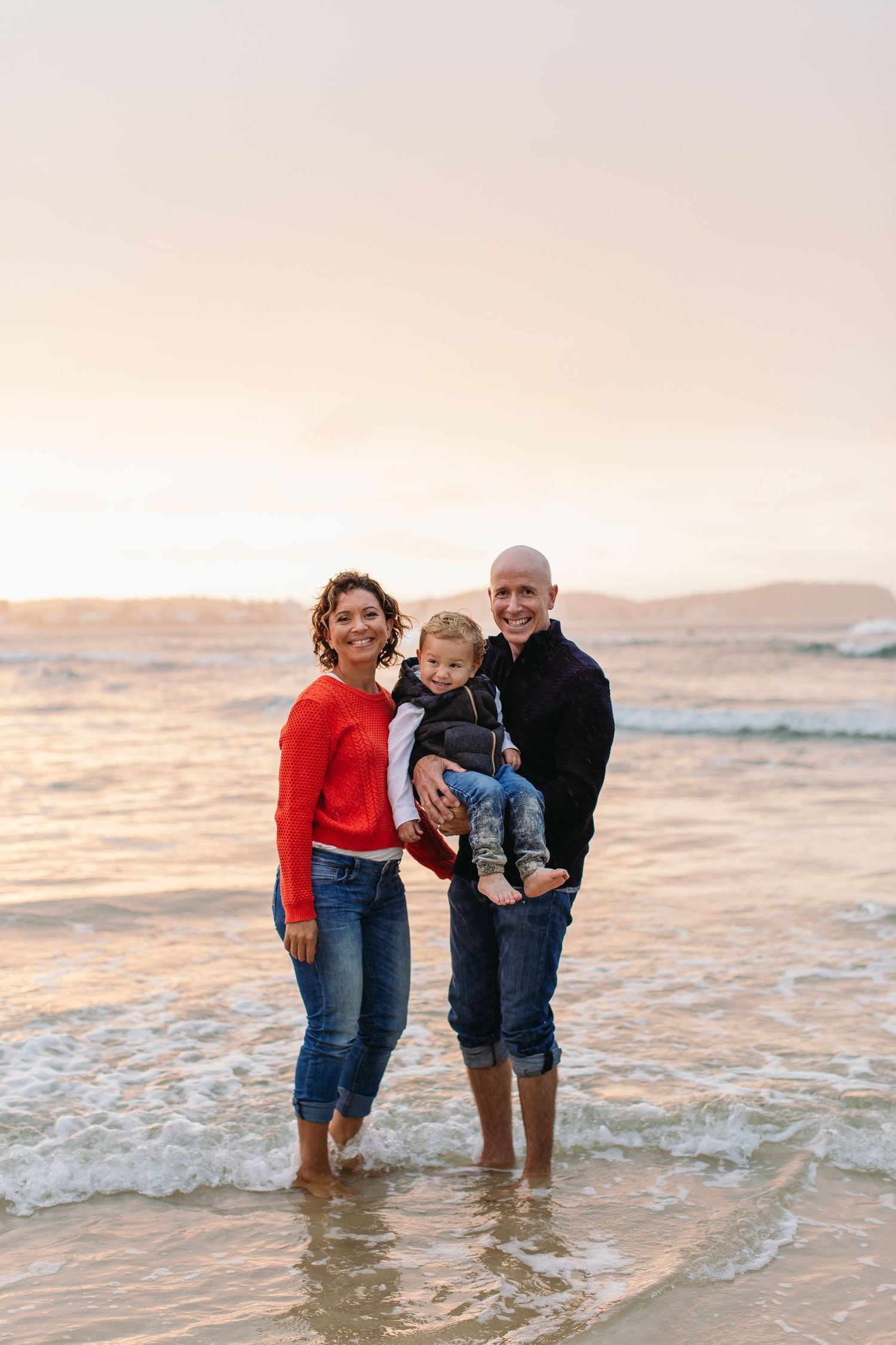Happy family of three standing barefoot in ocean waves at sunset, parents holding young child on beach.