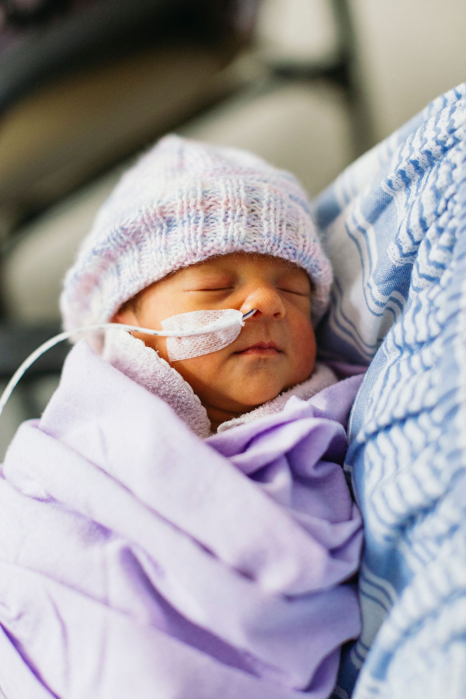 Newborn baby sleeping in hospital, swaddled in purple blanket with knit hat and nasal feeding tube.