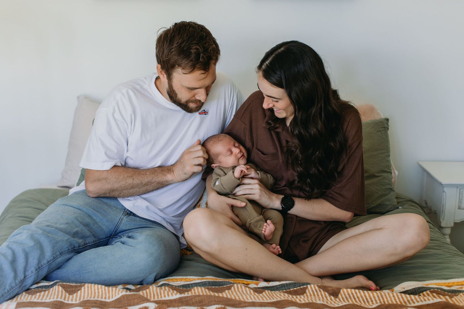 New parents sitting on bed together, lovingly cradling their newborn baby in a bright, cozy bedroom.