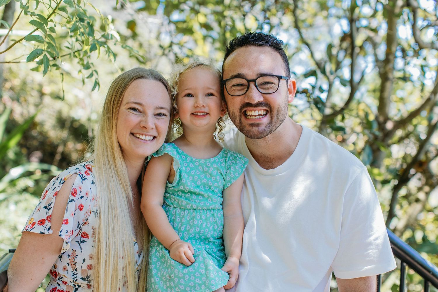 A smiling family of three poses outdoors, with parents holding a young blonde girl in a teal dress among green trees.