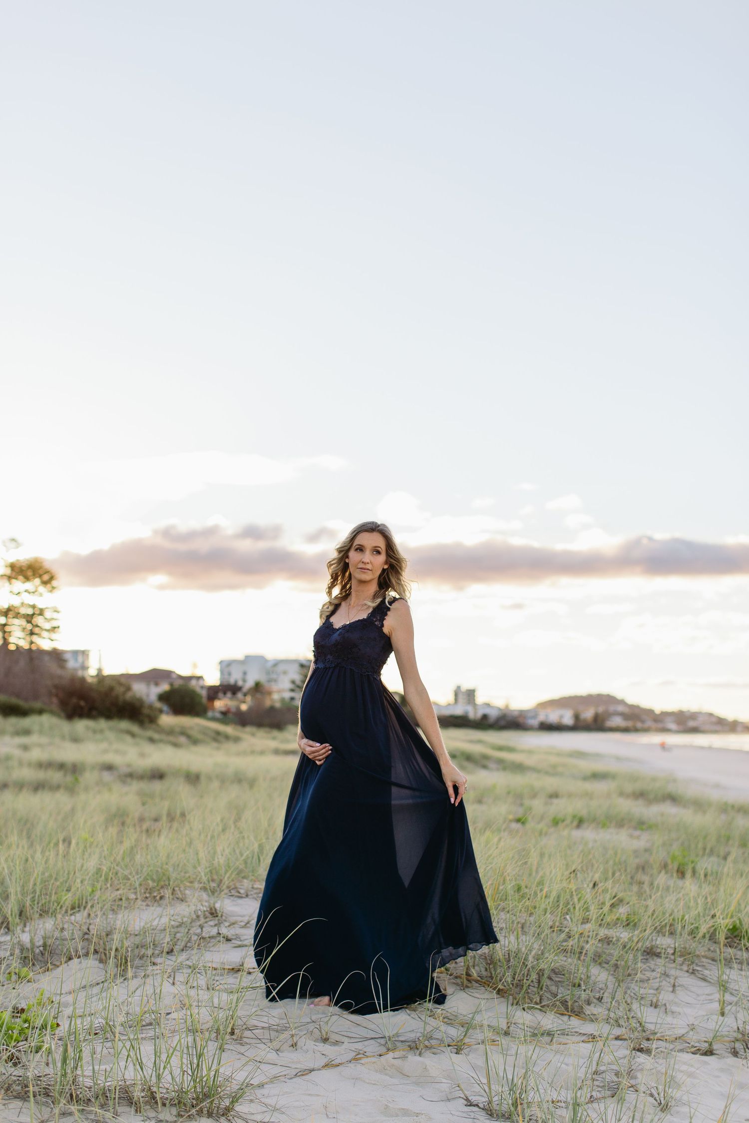 Pregnant woman in flowing navy blue dress standing on sandy beach dunes at sunset, cradling baby bump.