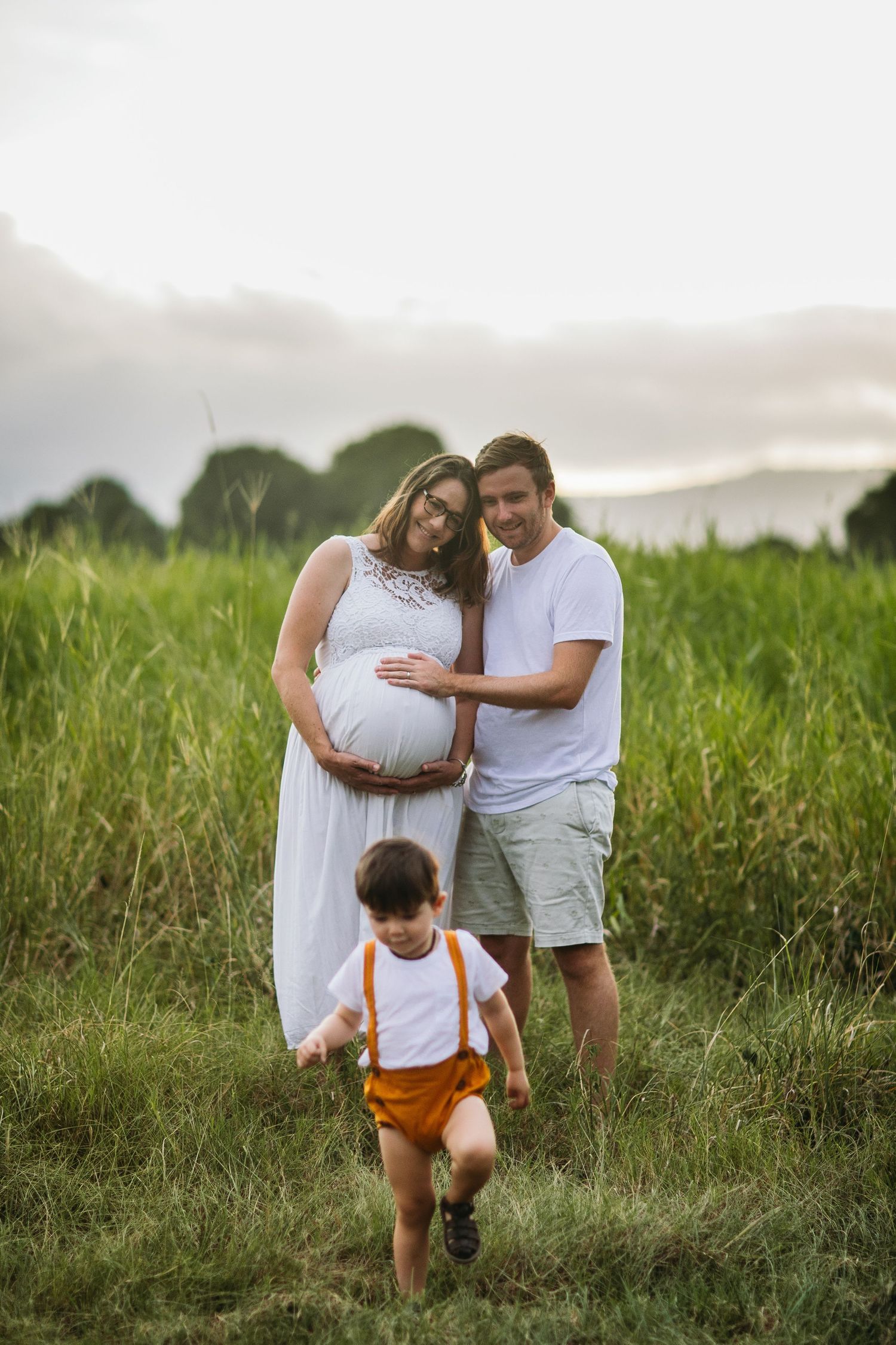 Pregnant woman in white dress with husband and toddler son in a lush green field during golden hour.