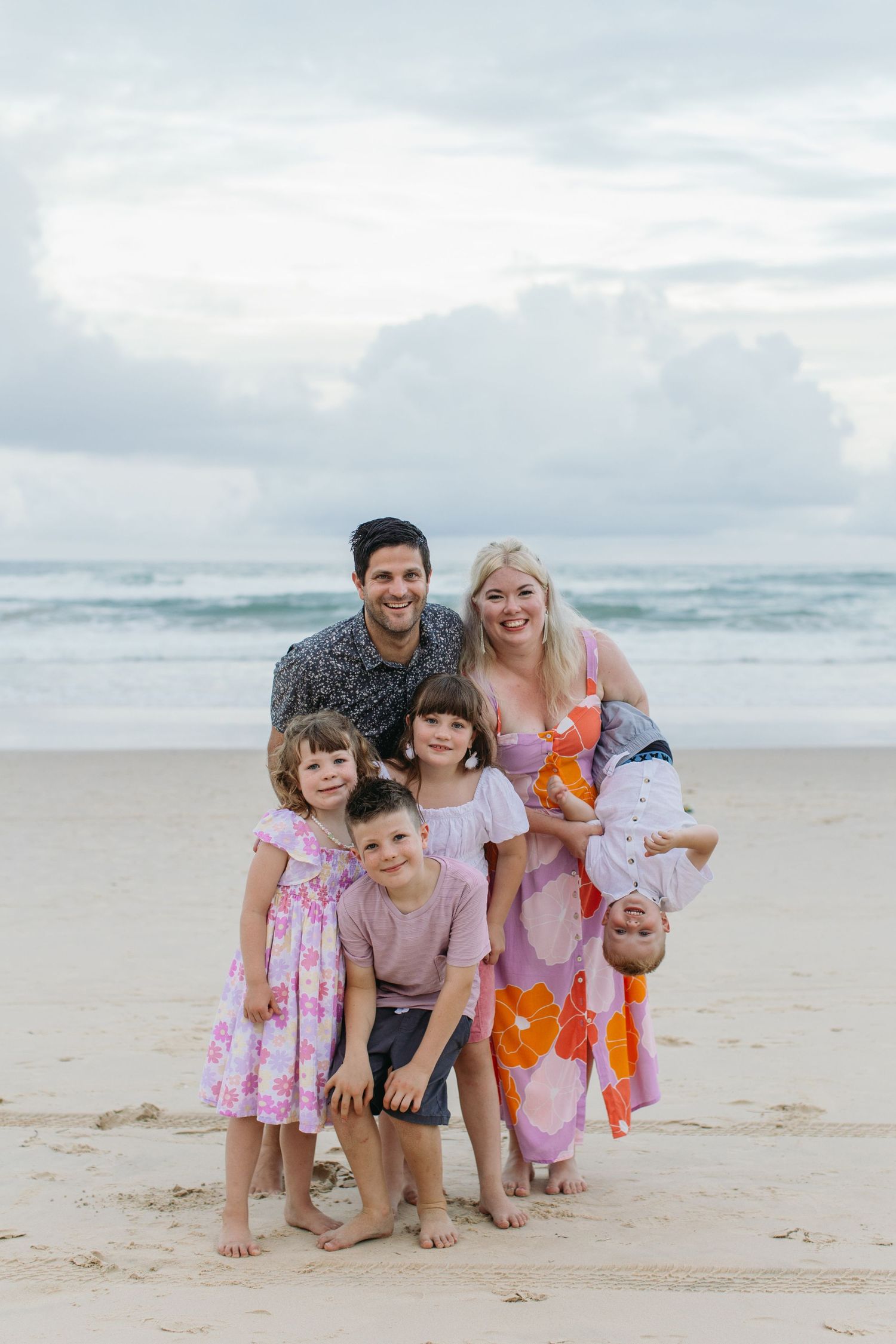 Smiling family of six standing barefoot on beach with waves and cloudy sky behind them.