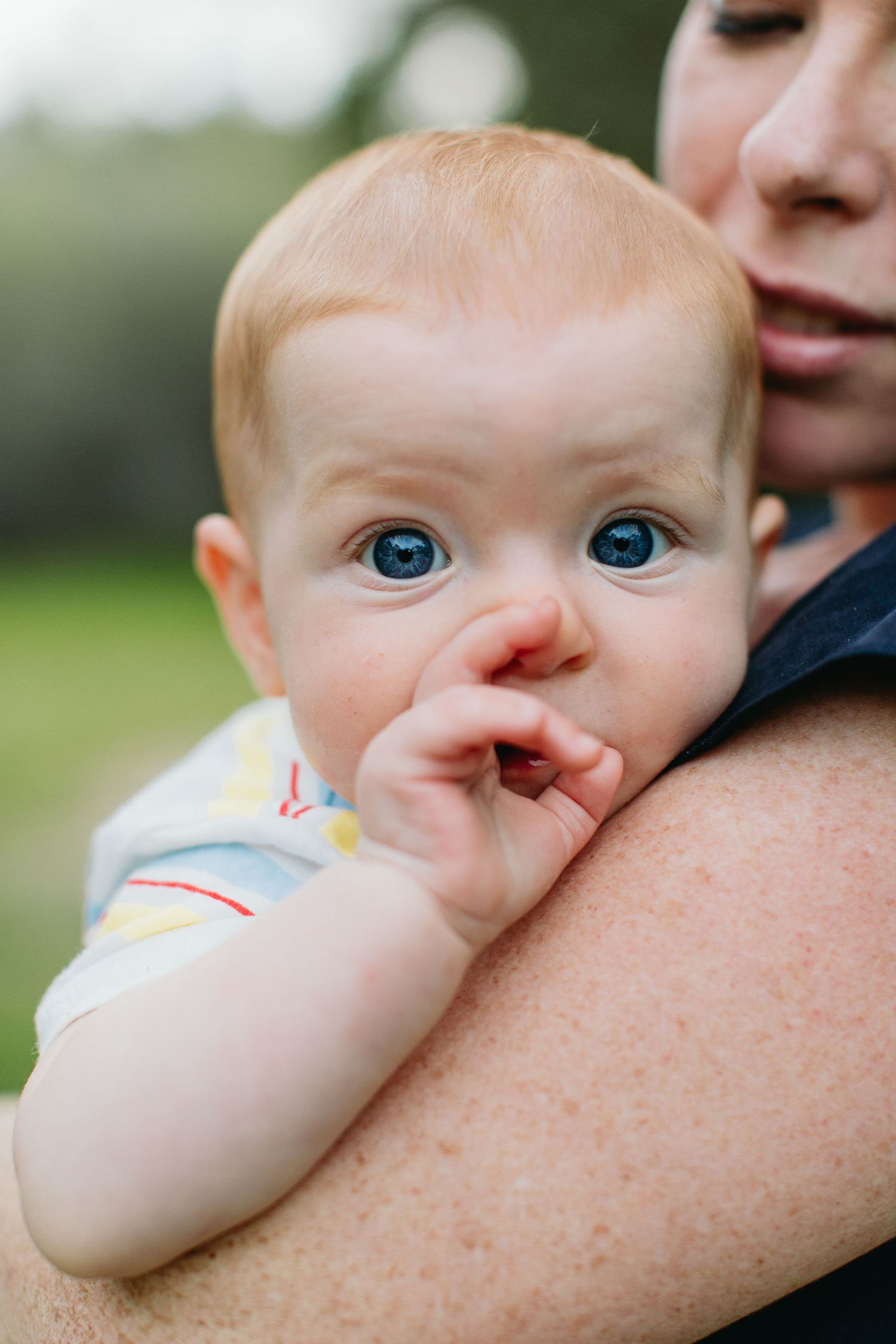 Adorable red-haired baby with blue eyes chewing fingers while being held by an adult outdoors.