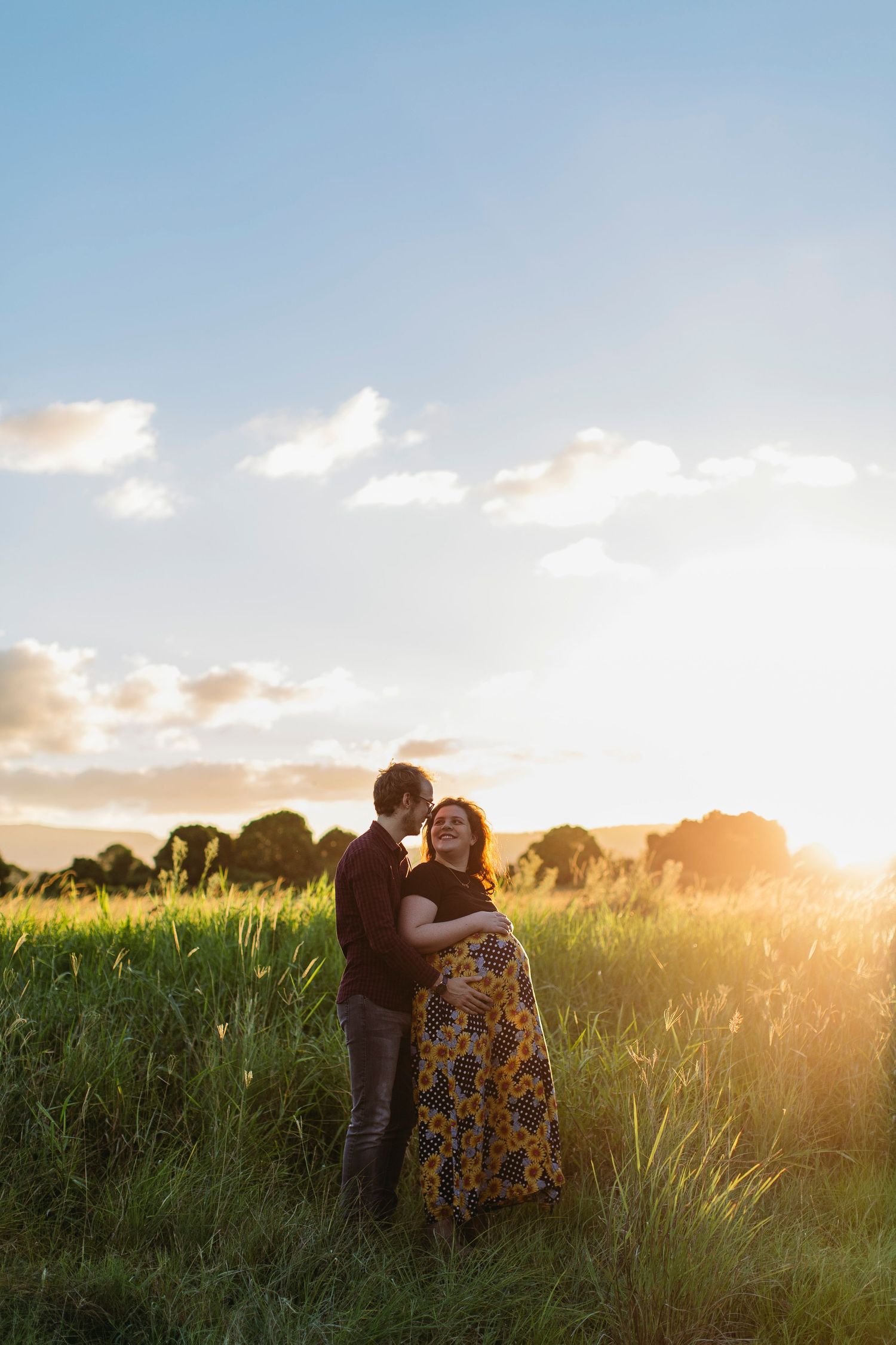Couple embracing in a sunlit field at golden hour, woman in floral maxi dress, lush green grass and blue sky backdrop.