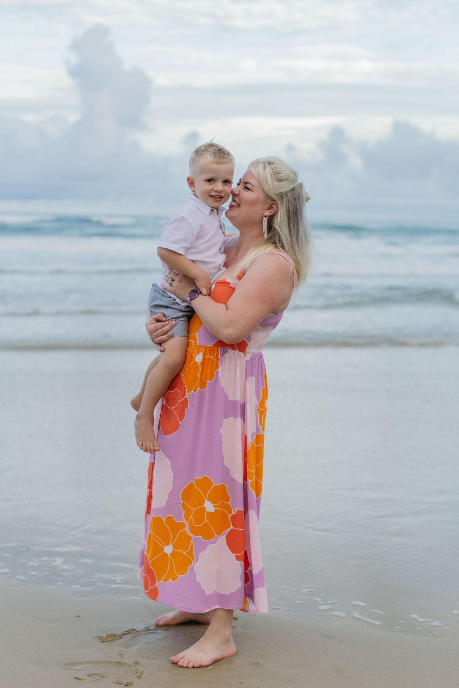 A smiling mother in a colorful floral dress cuddles her laughing toddler son on a sandy beach.