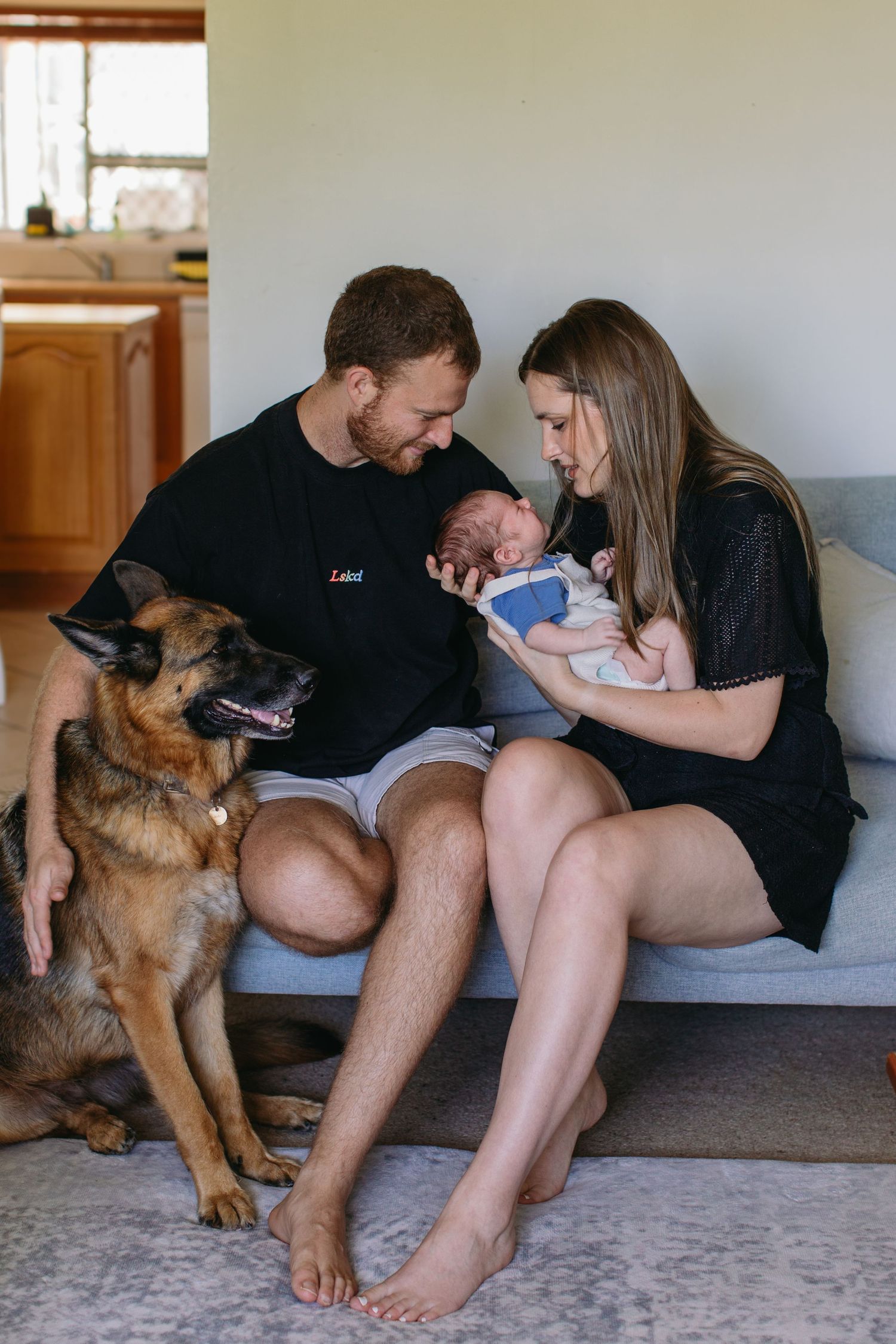 Young family with newborn baby and German Shepherd dog sitting together on couch at home.