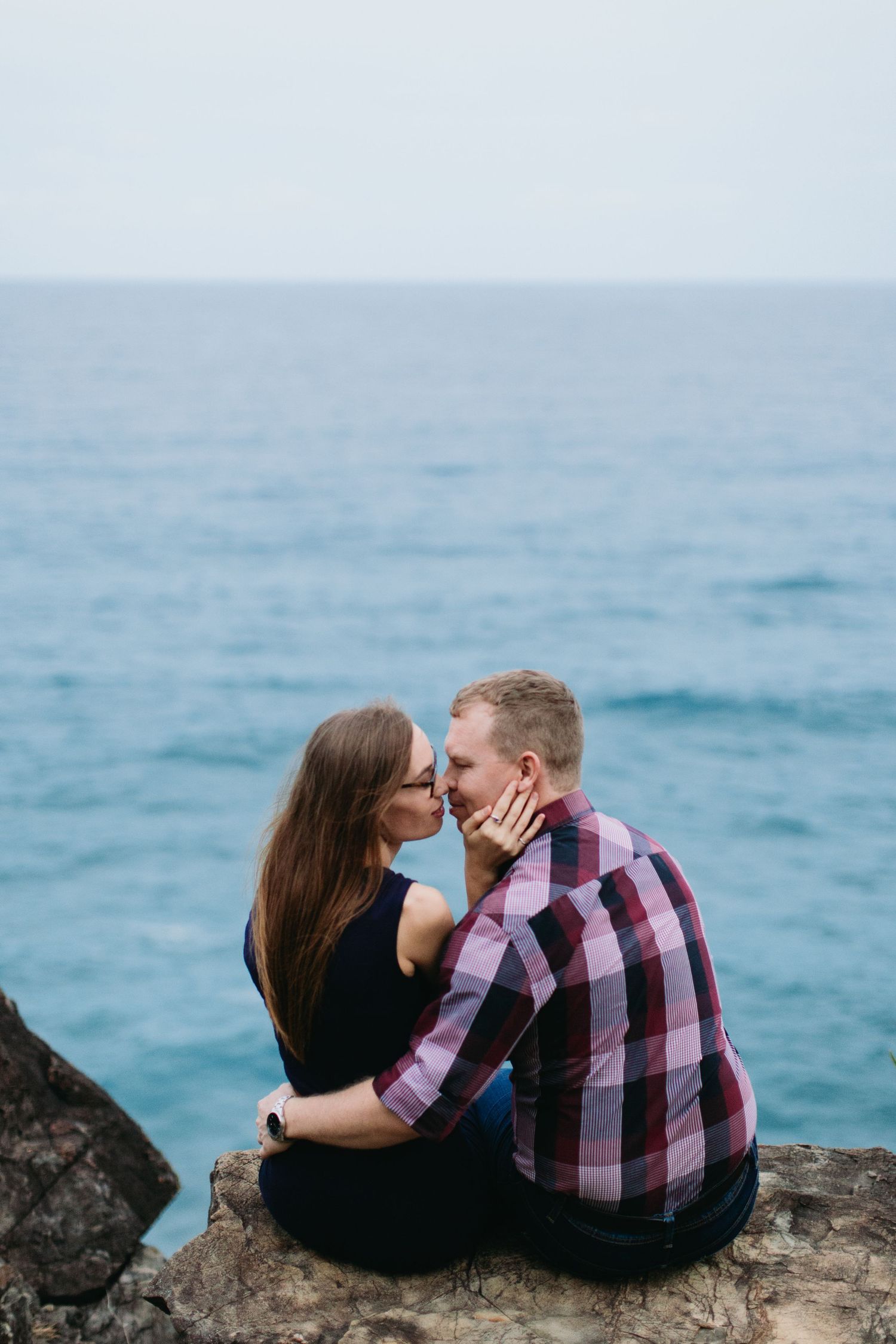 Couple sharing a romantic kiss while sitting on rocky cliffs overlooking a serene blue ocean.