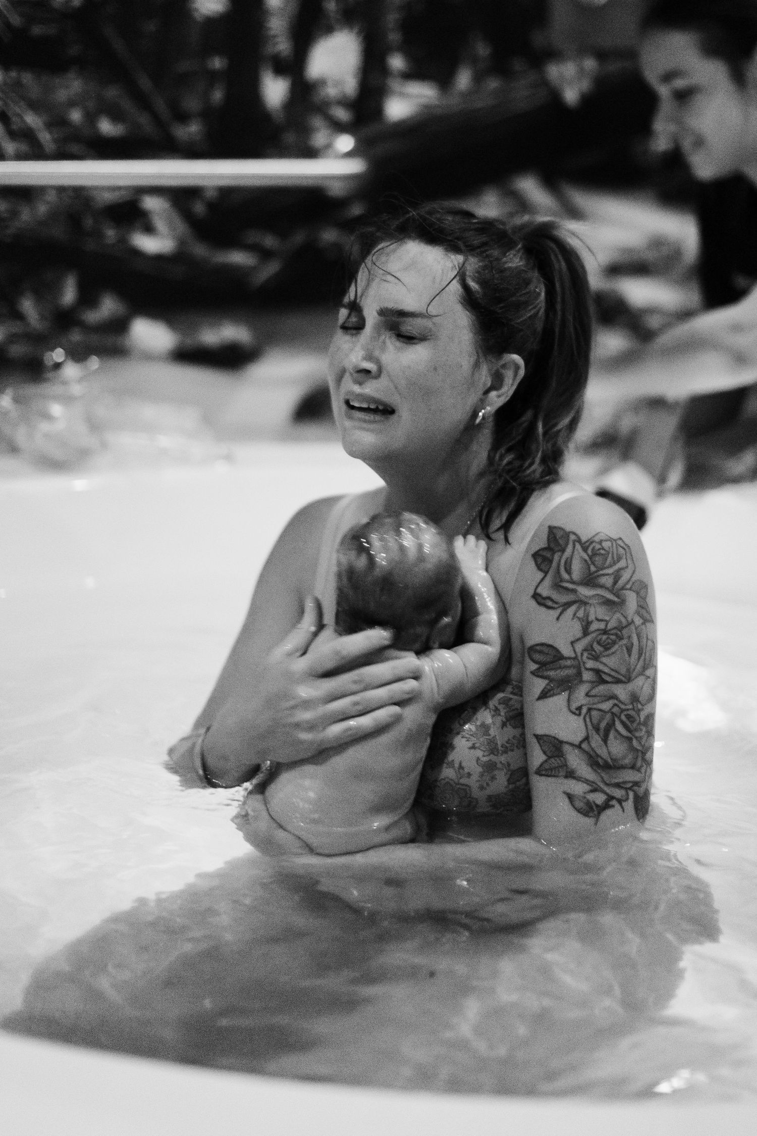 Emotional mother holding newborn during water birth, black and white photo with rose tattoo visible.