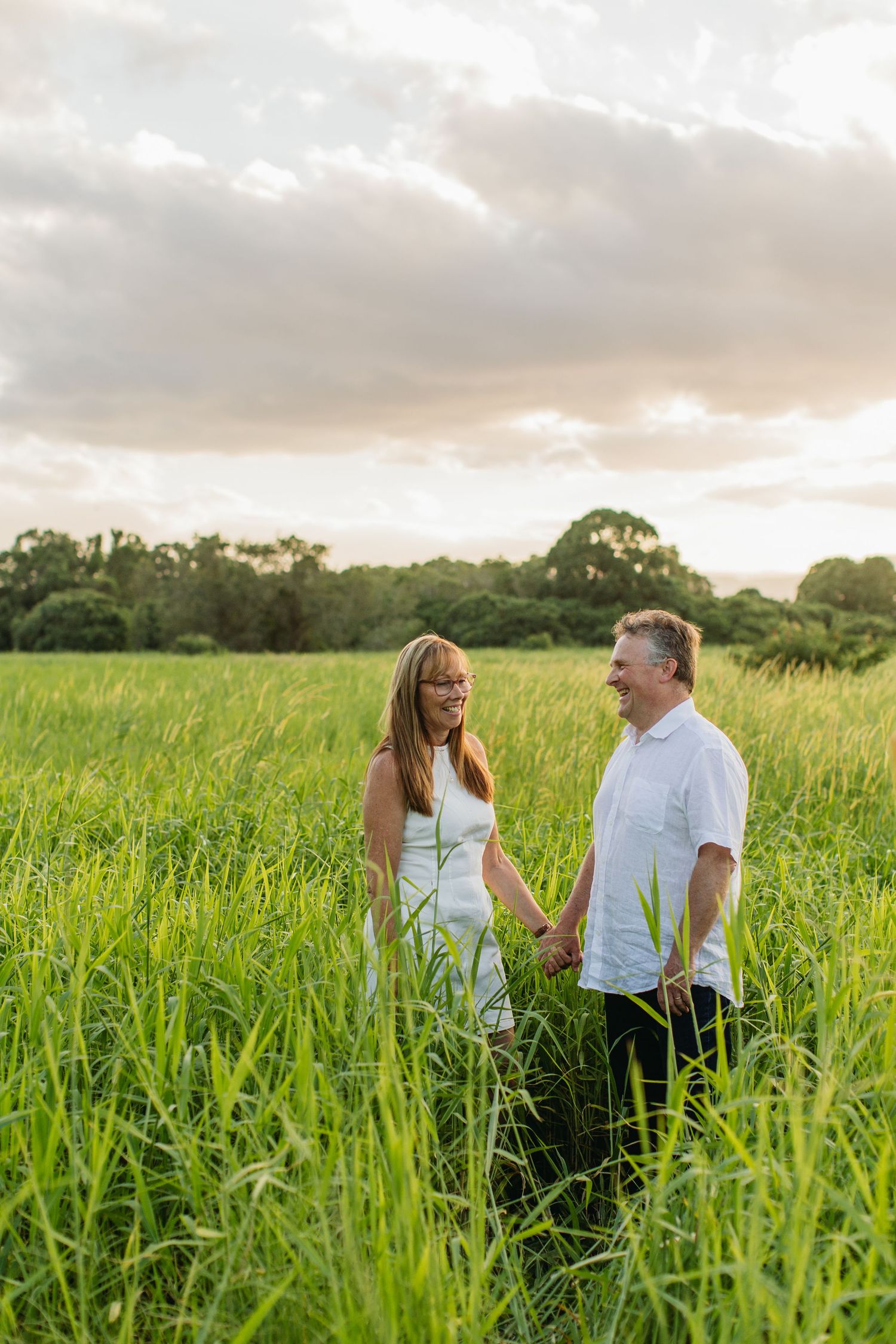 Couple holding hands smiling at each other in a lush green field at sunset wearing white outfits.