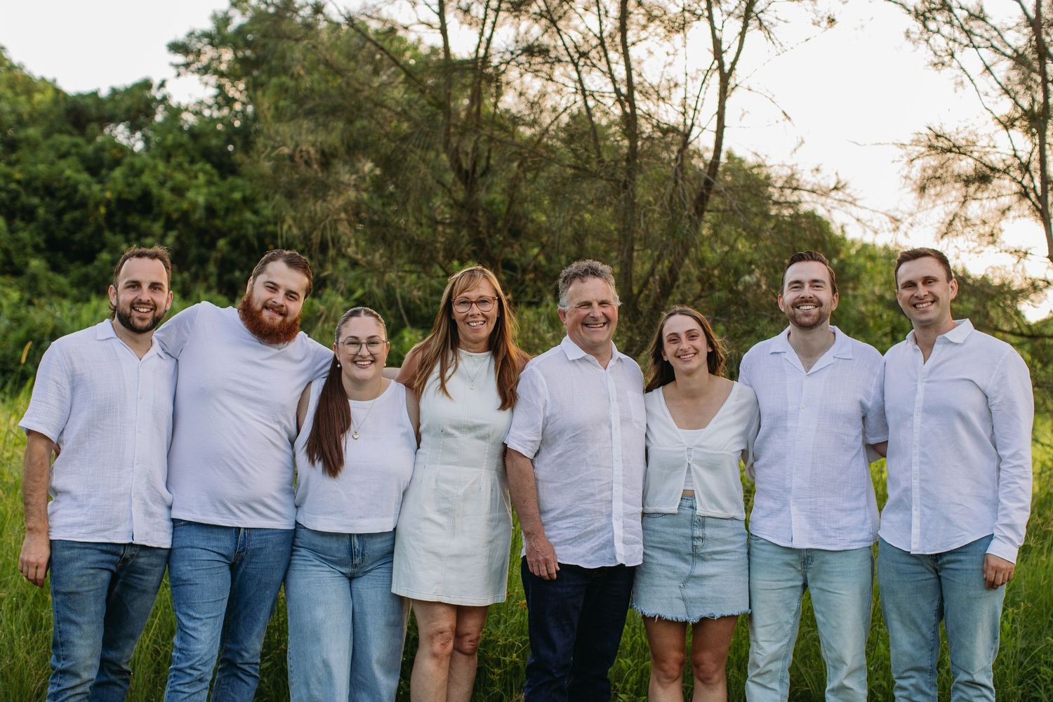 A joyful family of eight poses together outdoors in matching white shirts and jeans, surrounded by lush green trees.