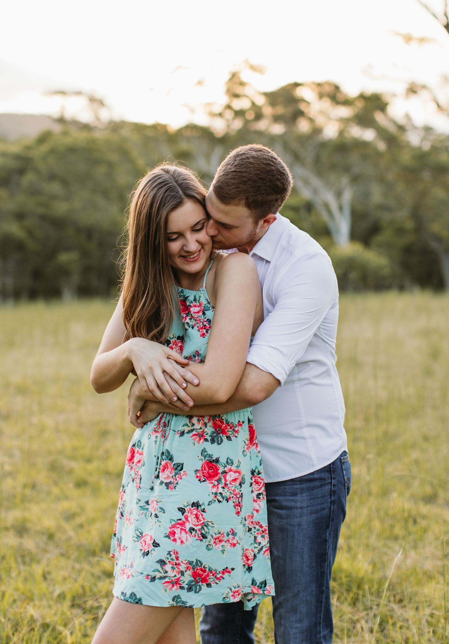 Couple embracing in a field, woman in floral dress smiling as man kisses her cheek at golden hour.