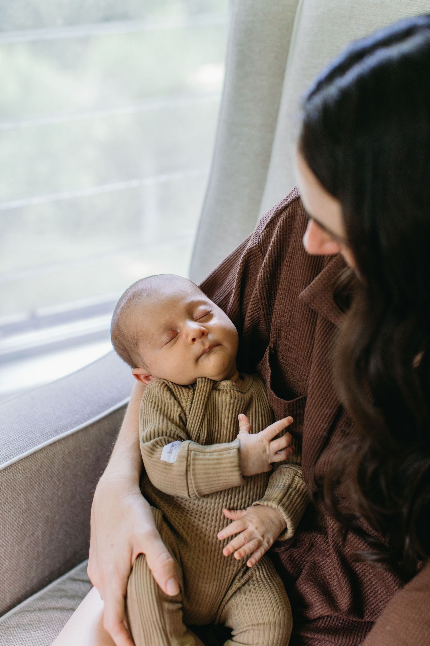 A mother in a brown cardigan tenderly holds her sleeping newborn baby dressed in an olive green ribbed onesie.