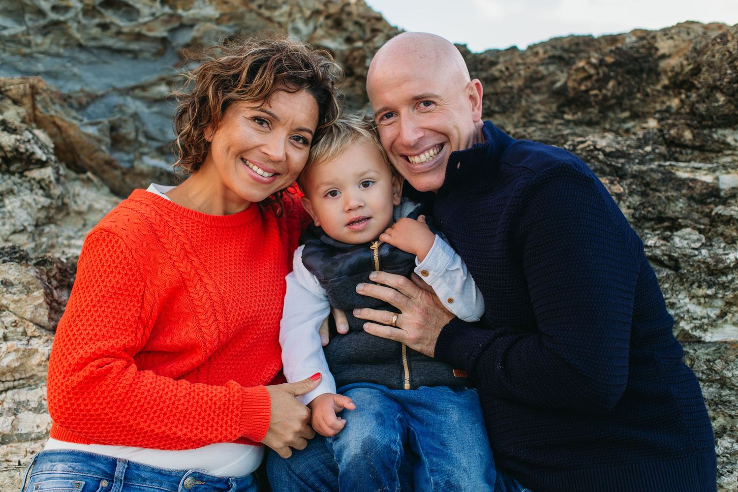 Happy family of three posing outdoors near rocky cliffs, parents in red and navy sweaters smiling with toddler.