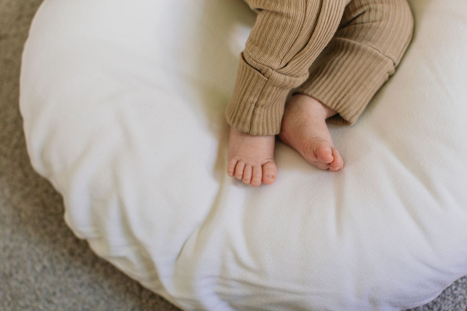 Close-up of baby feet resting on a white nursing pillow, wearing brown ribbed pants.