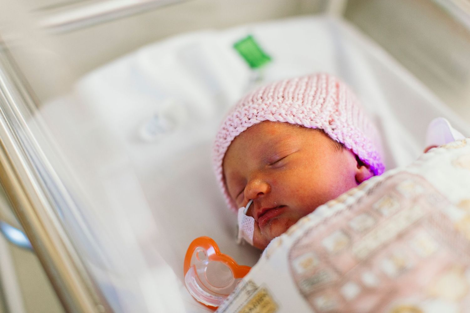 Newborn baby sleeping in hospital bassinet wearing pink knit hat with pacifier and nasal feeding tube.