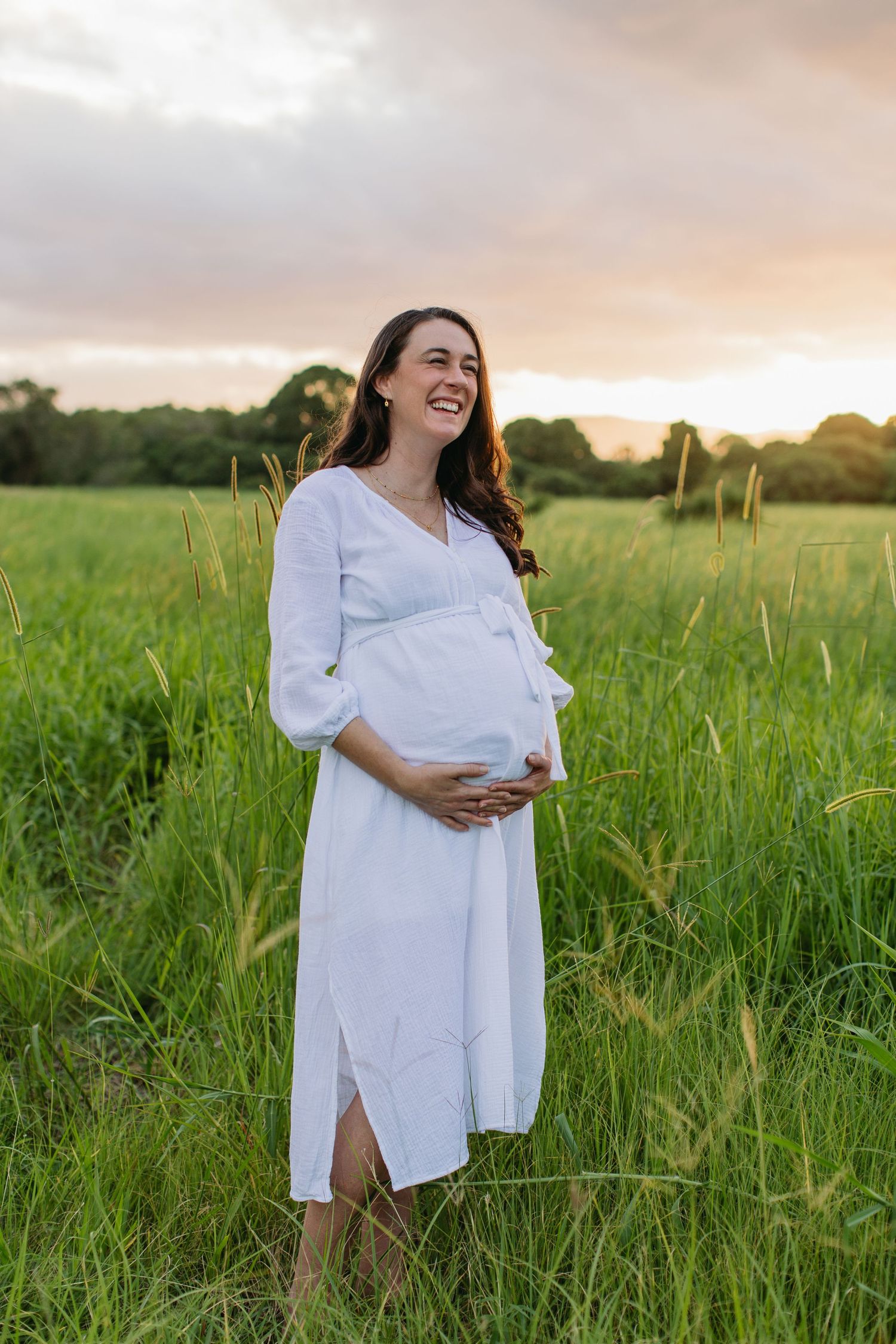 Smiling pregnant woman in white dress holding belly in lush green field at sunset.