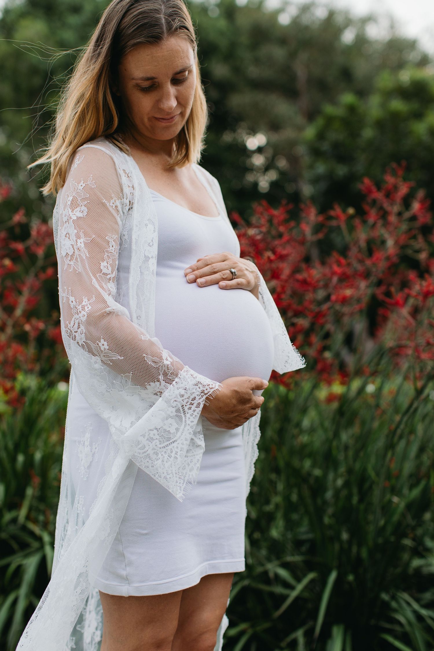 Pregnant woman in white lace robe cradling baby bump outdoors surrounded by lush red and green garden foliage.