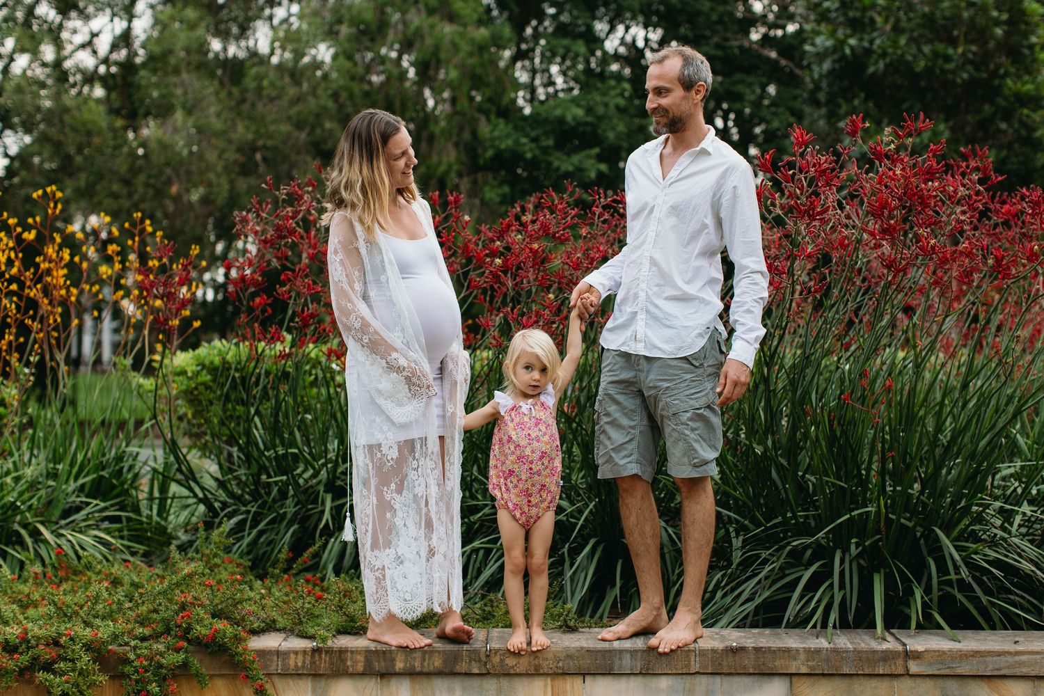 Pregnant woman in white lace dress with man and toddler girl holding hands in a colorful garden.