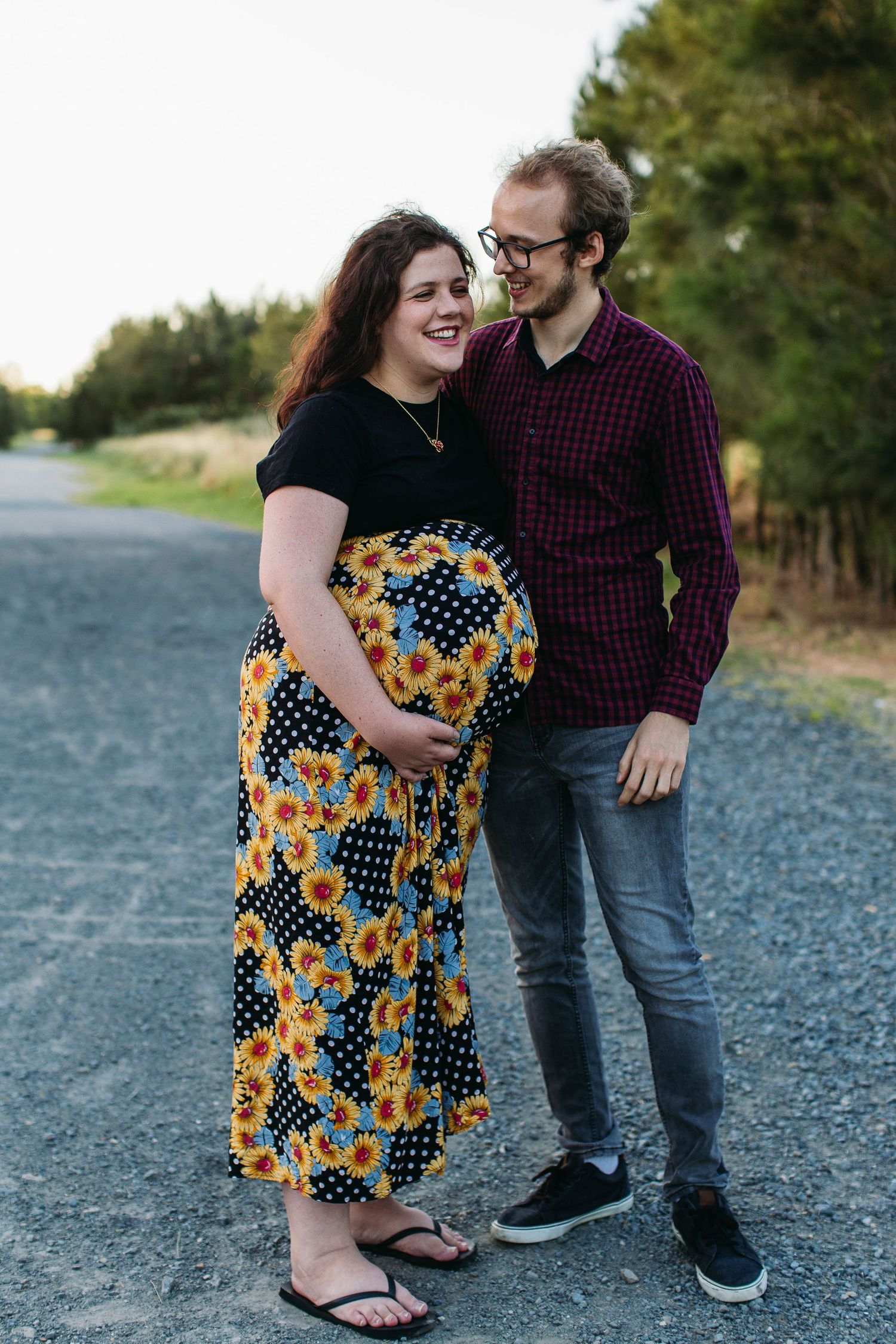 Pregnant woman in sunflower skirt and man in plaid shirt share a tender moment on a rural road.