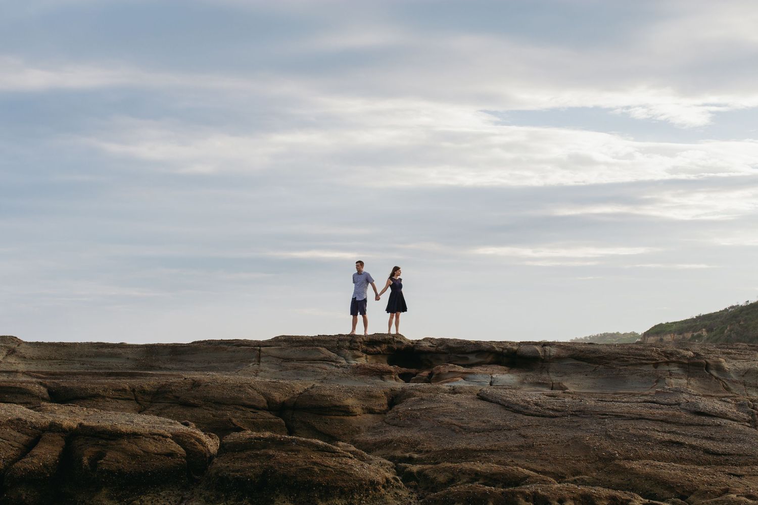 Couple holding hands standing on rocky coastal cliffs with a dramatic cloudy sky backdrop at sunset.