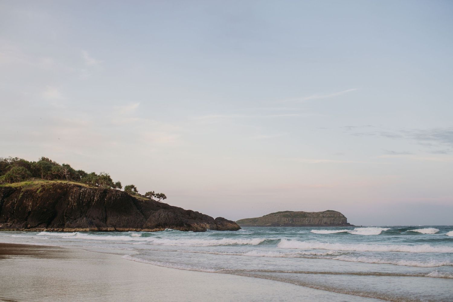 Serene coastal beach scene with gentle waves, rocky headland, and soft pastel sky at dusk.