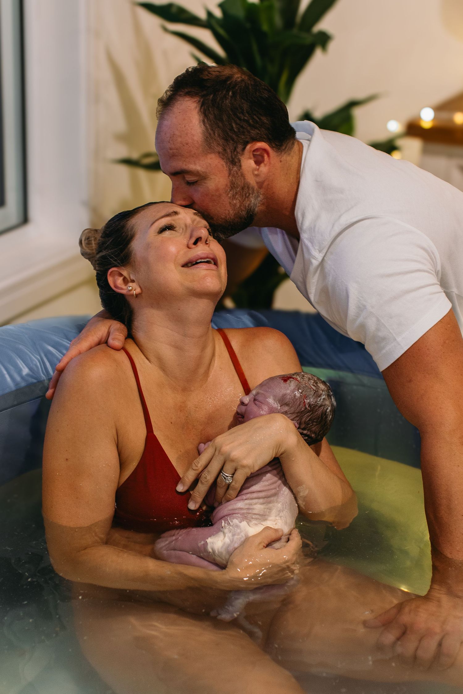 Emotional new mother cradles newborn in birth pool as partner kisses her head during water birth.