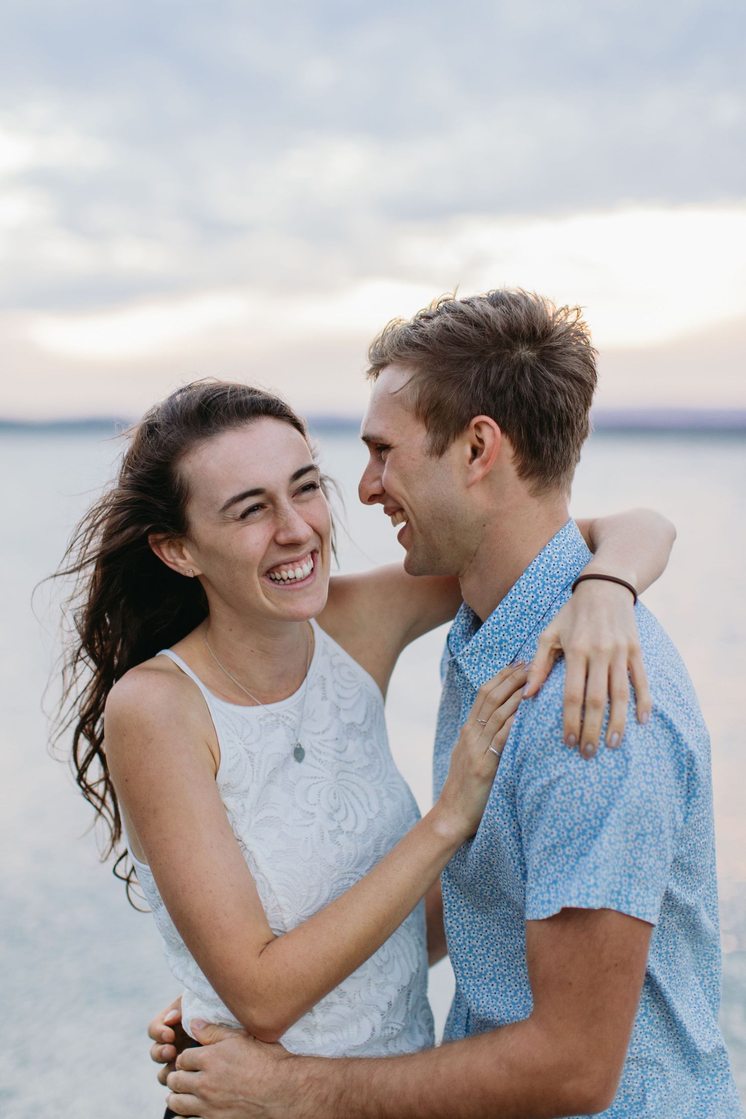 Happy couple embracing and laughing together on a beach at sunset, wearing casual summer clothing.