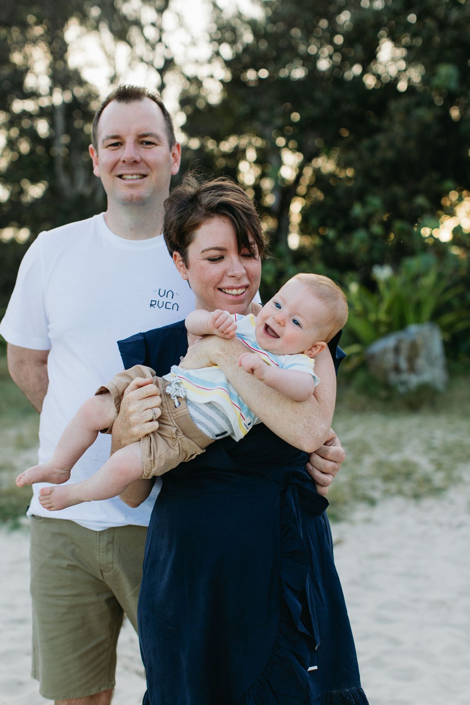 Happy young family with mother holding smiling baby on a sandy beach, father standing beside them outdoors.