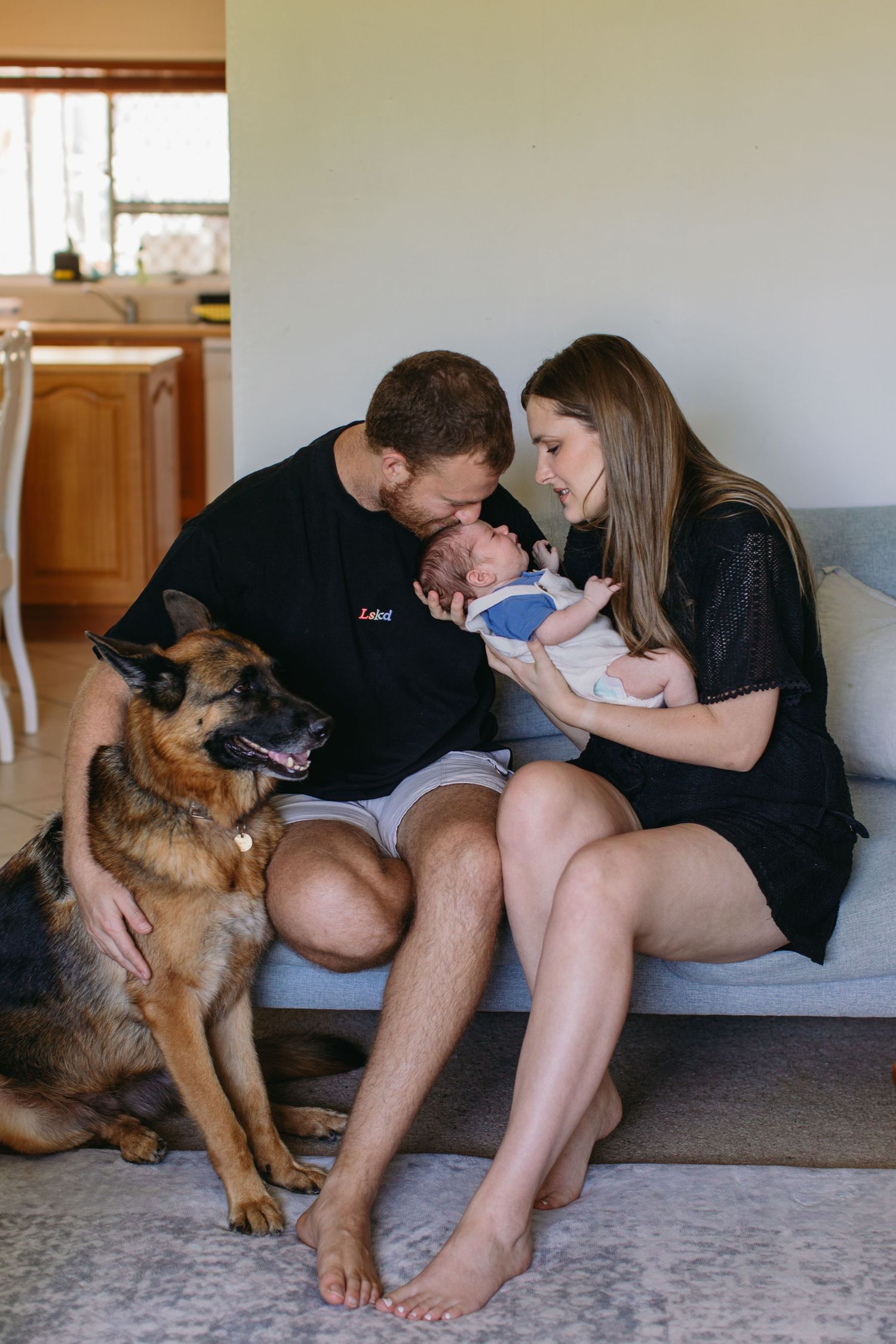 Happy parents kissing newborn baby on sofa with German Shepherd dog sitting beside them.