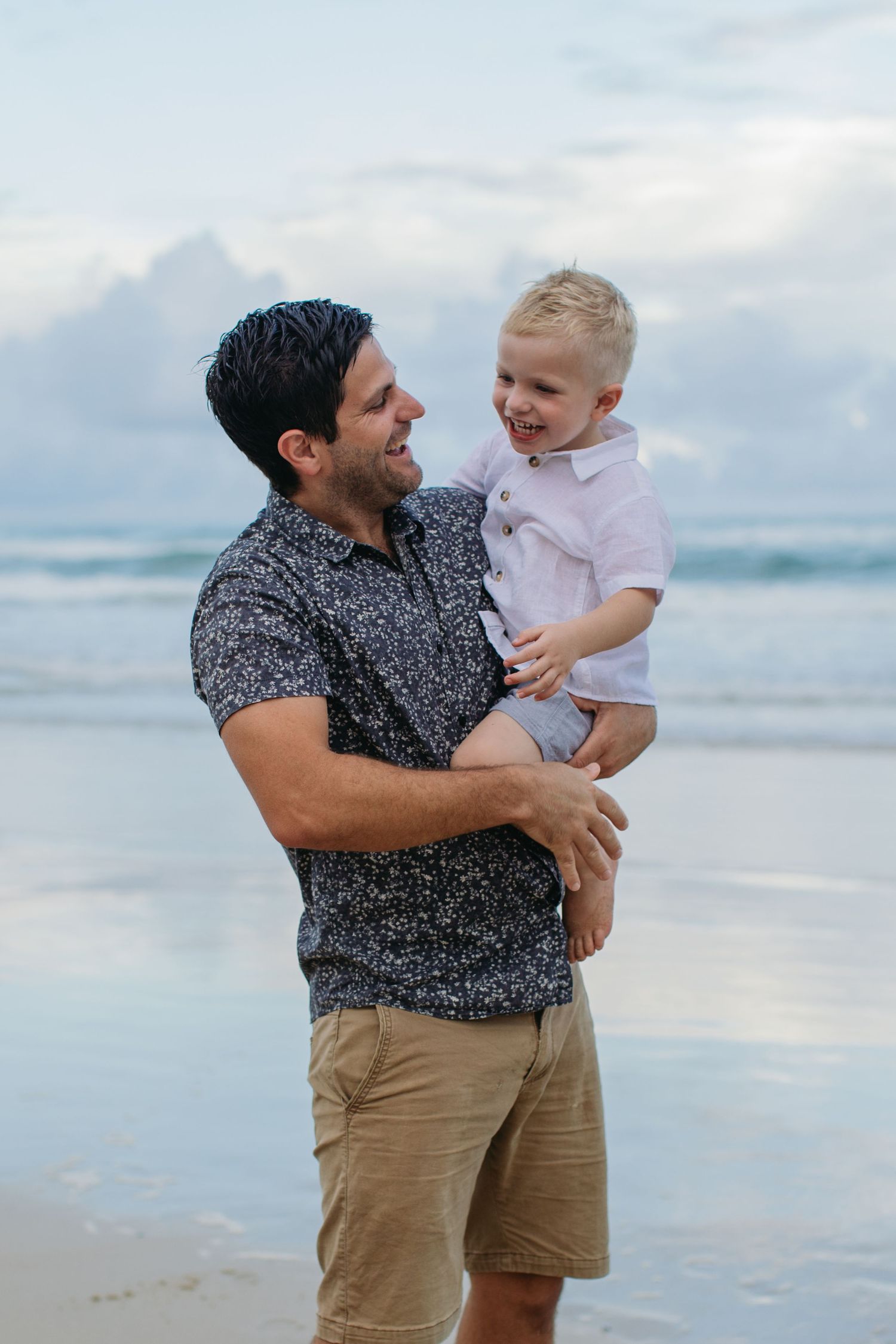 A smiling father holds his laughing blonde toddler son on a beach with ocean waves in the background.