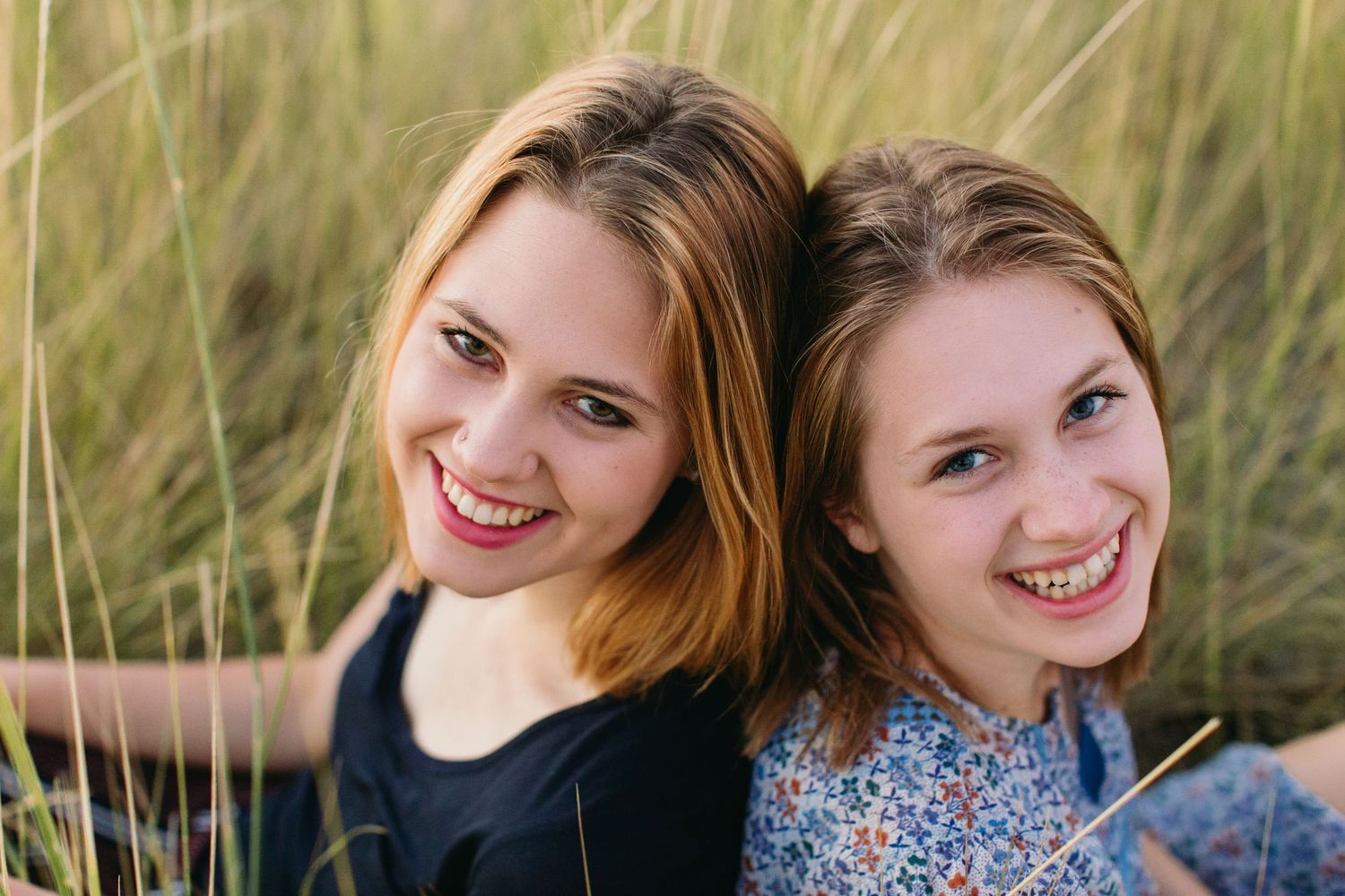 Two smiling young women sitting back-to-back outdoors in a grassy field, enjoying a sunny day together.