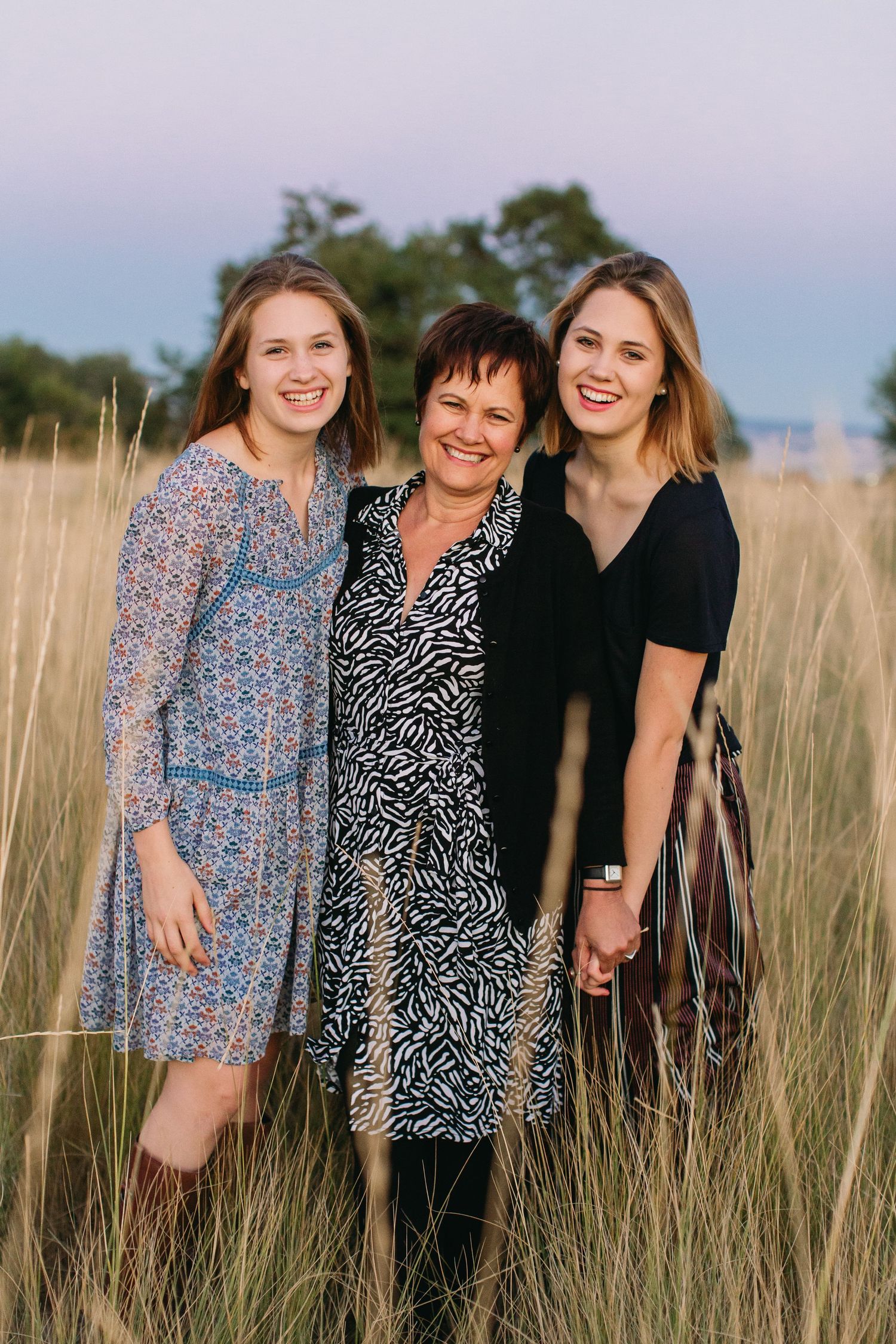 Three women smiling together in a golden grass field at dusk, wearing floral and patterned dresses.