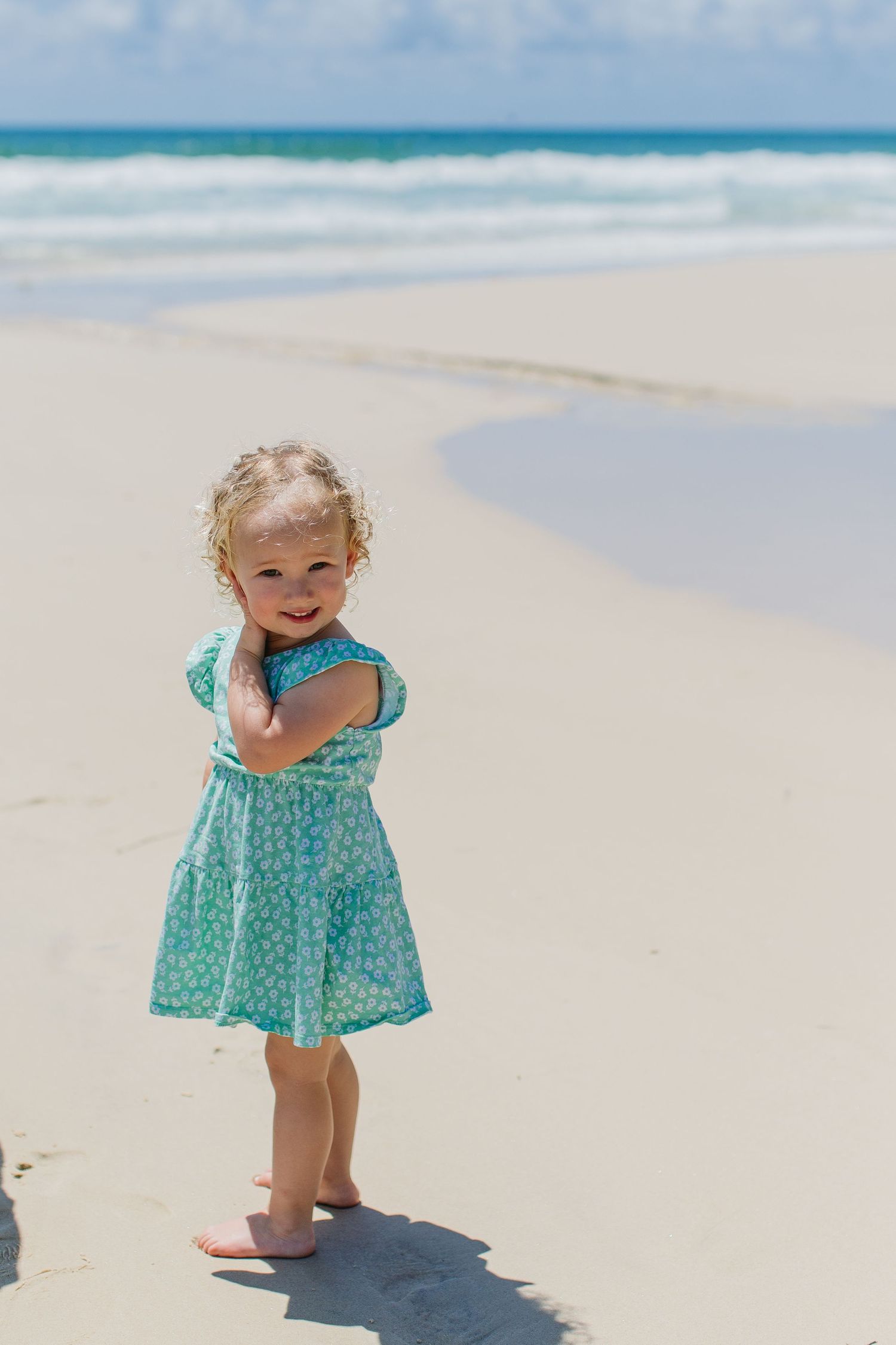 Adorable toddler girl in a teal floral dress standing barefoot on a sunny sandy beach with ocean waves behind her.