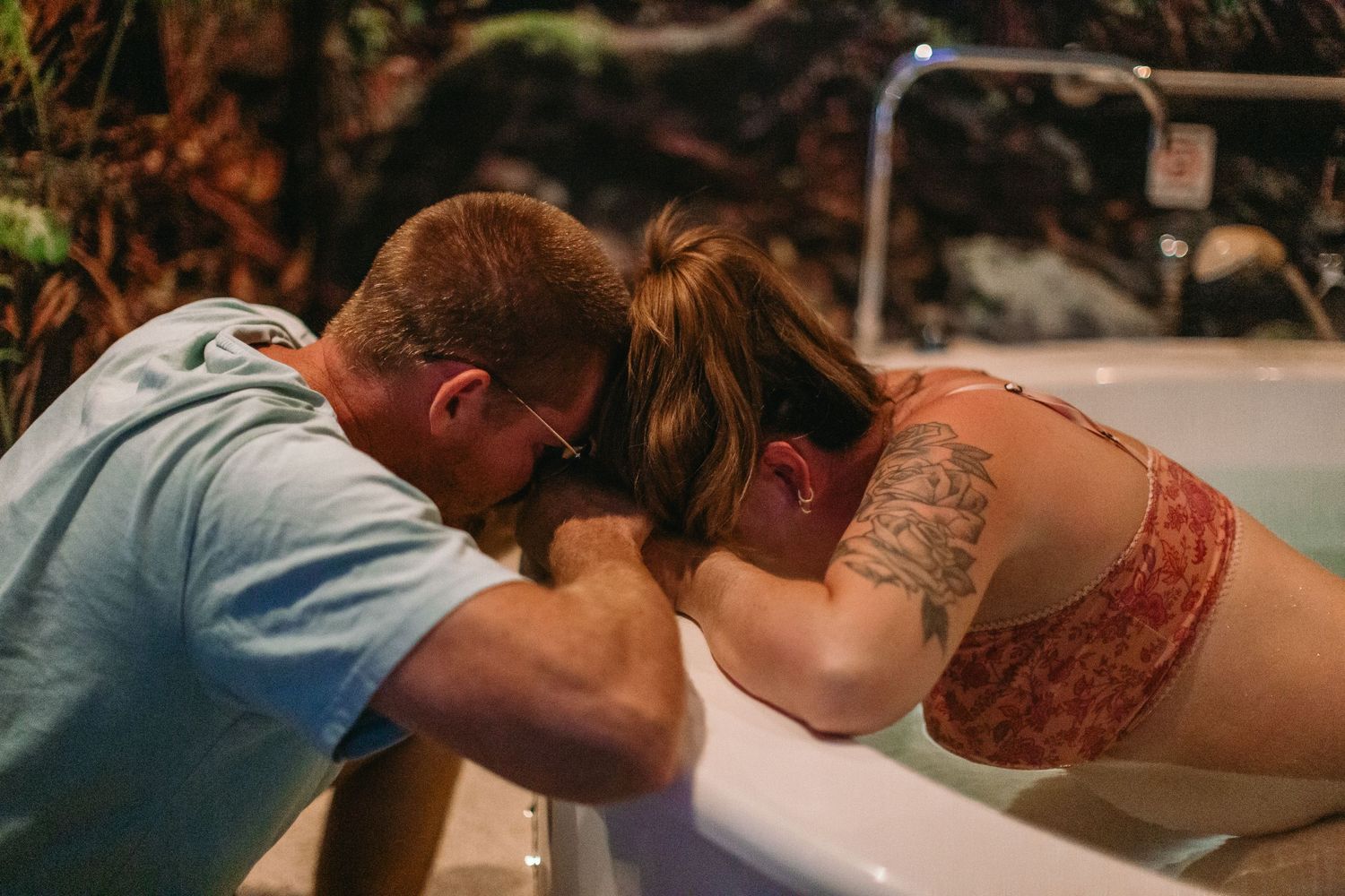 A couple shares an intimate moment leaning over a birthing tub during a water birth labor support session.