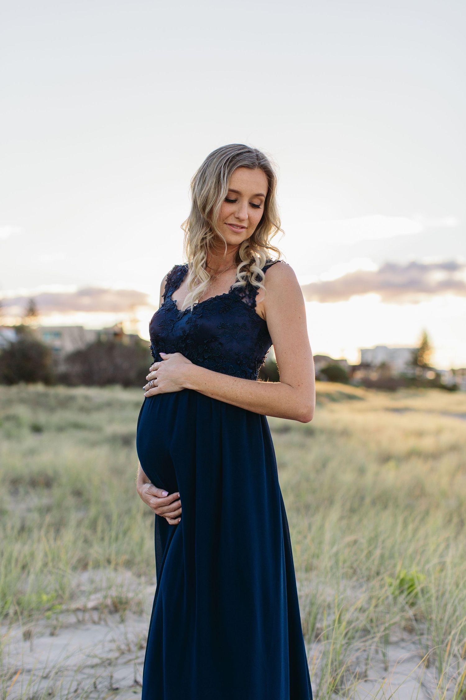 Pregnant woman in navy lace maternity gown cradling her baby bump outdoors on sandy dunes at sunset.