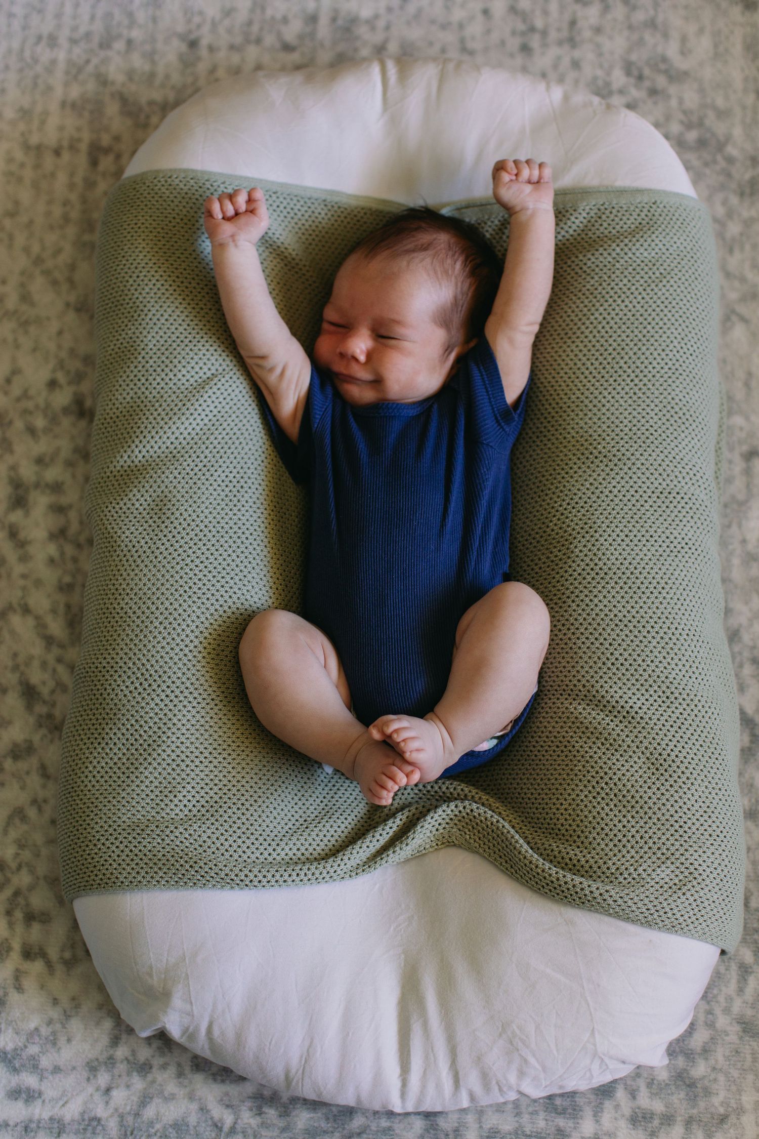 Newborn baby in blue onesie stretching arms up while lying in green and white nursing pillow.