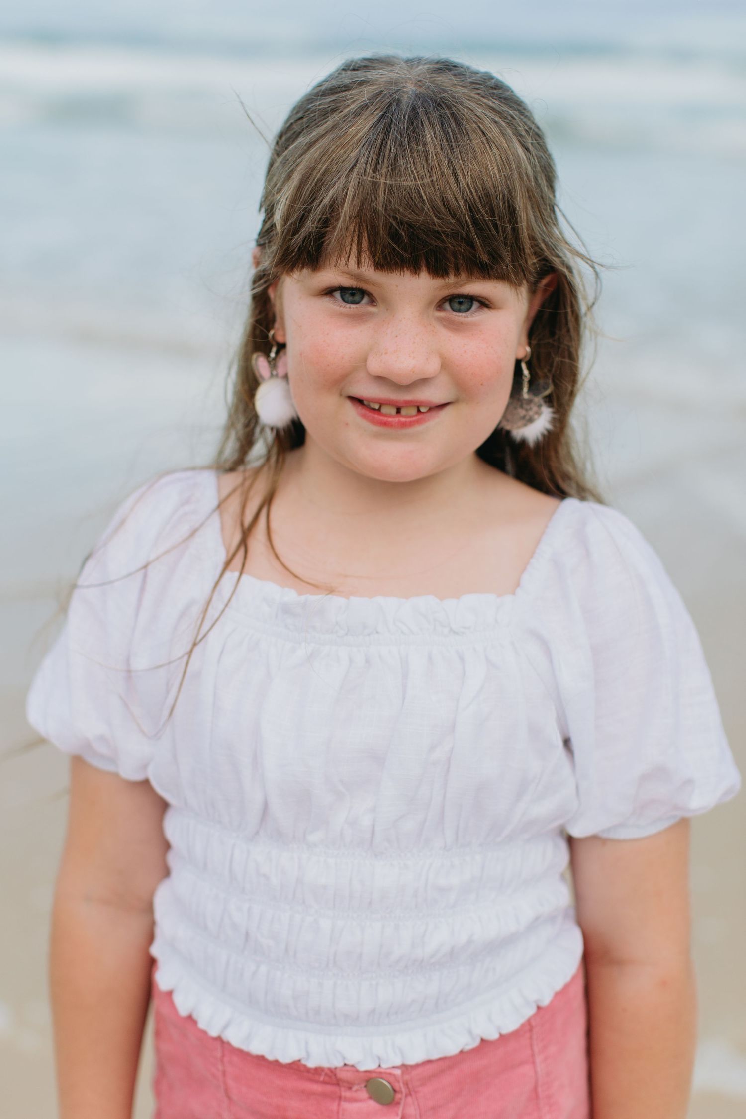 Young girl with bangs wearing a white puff-sleeve top and shell earrings smiling at the beach.