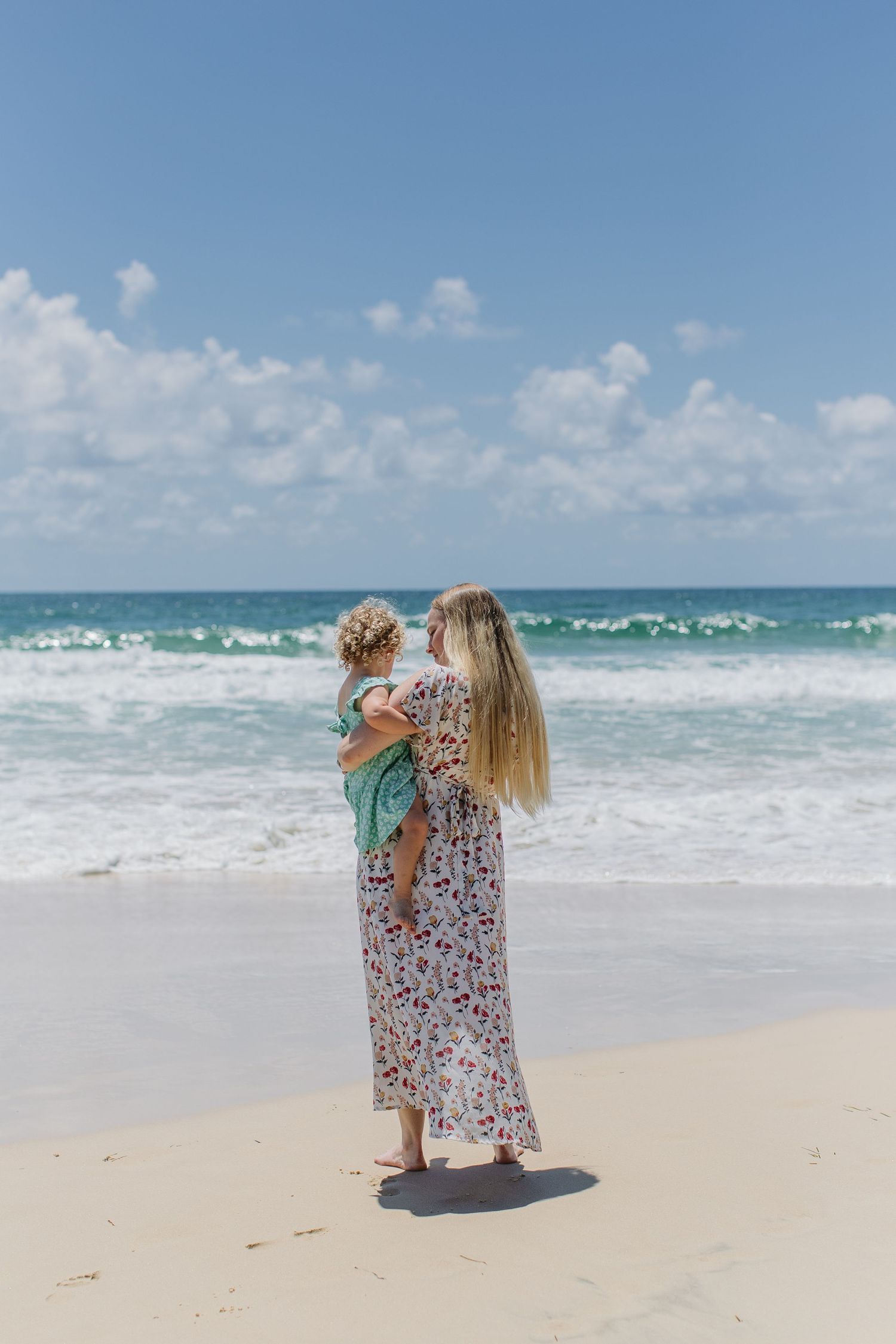 Mother in floral dress holding toddler facing turquoise ocean waves on sunny beach.