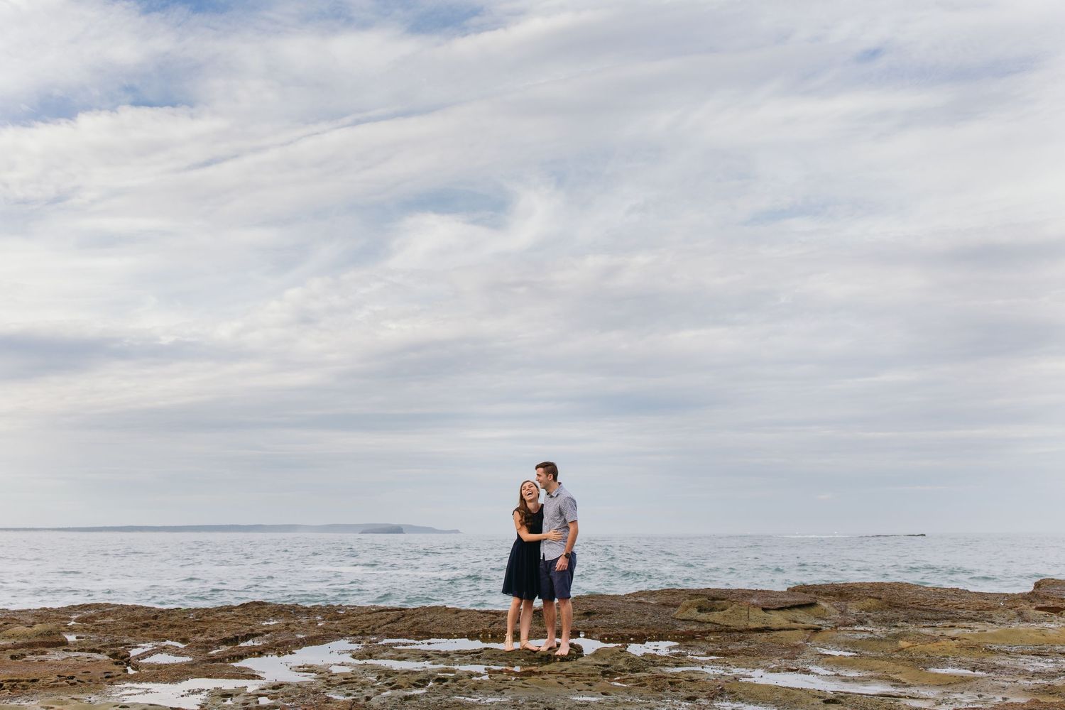 Couple embracing on rocky coastal shoreline with calm ocean and cloudy sky backdrop.