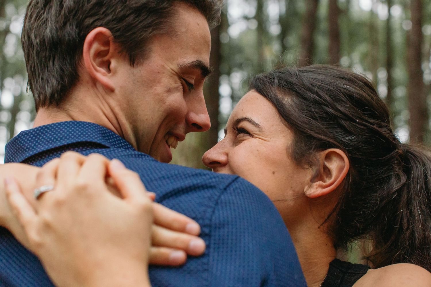A couple embraces and smiles at each other tenderly in a forest setting, the woman wearing a ring.