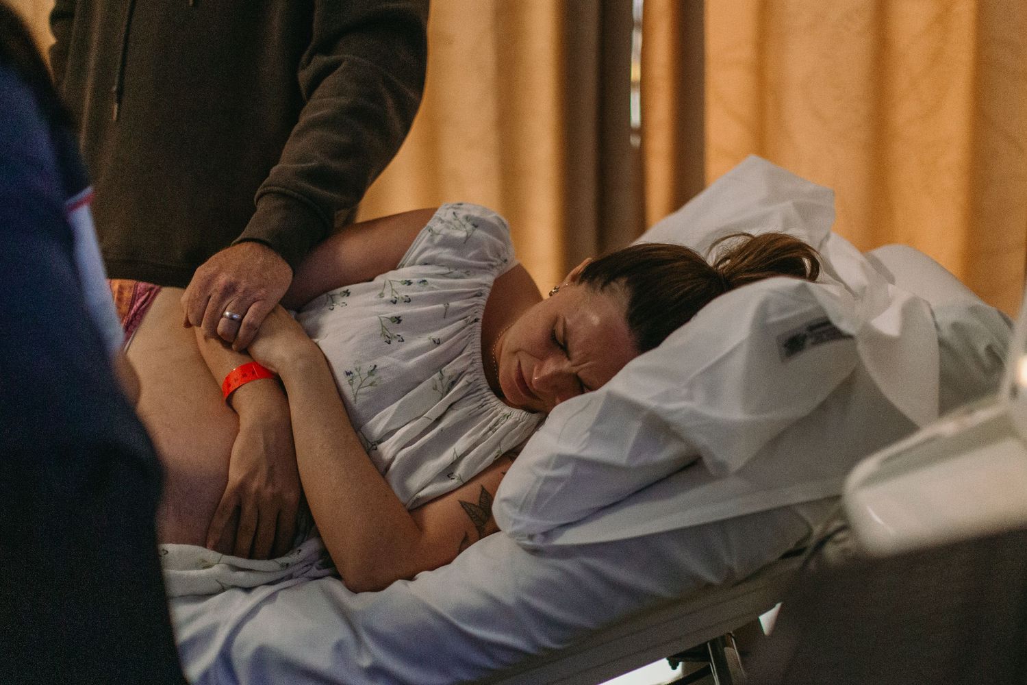 Pregnant woman resting in hospital bed during labor, supported by a partner standing beside her.